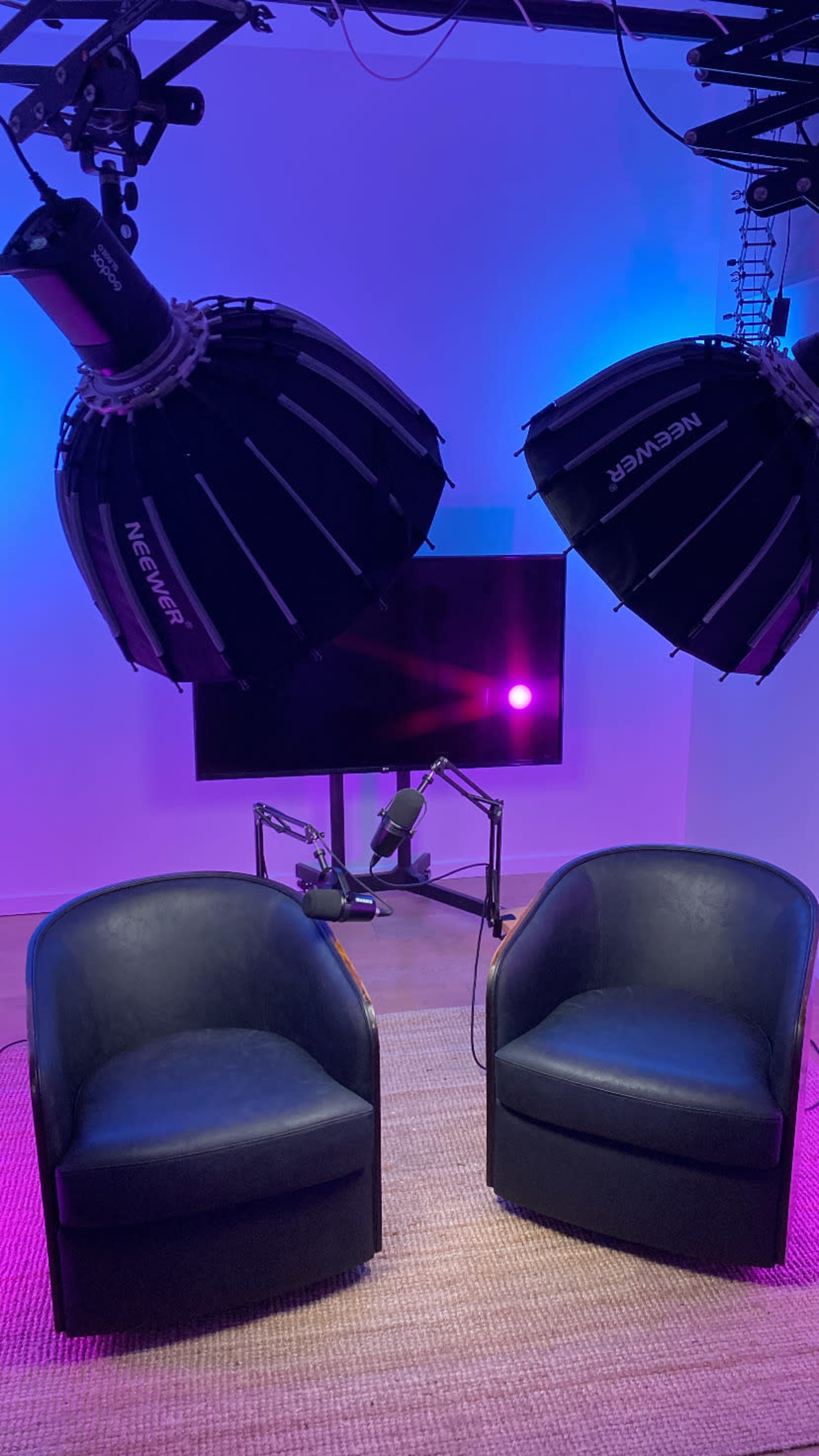 The image shows two dark blue armchairs positioned facing a television screen, with large studio lights overhead illuminating the scene in a well-lit setting.