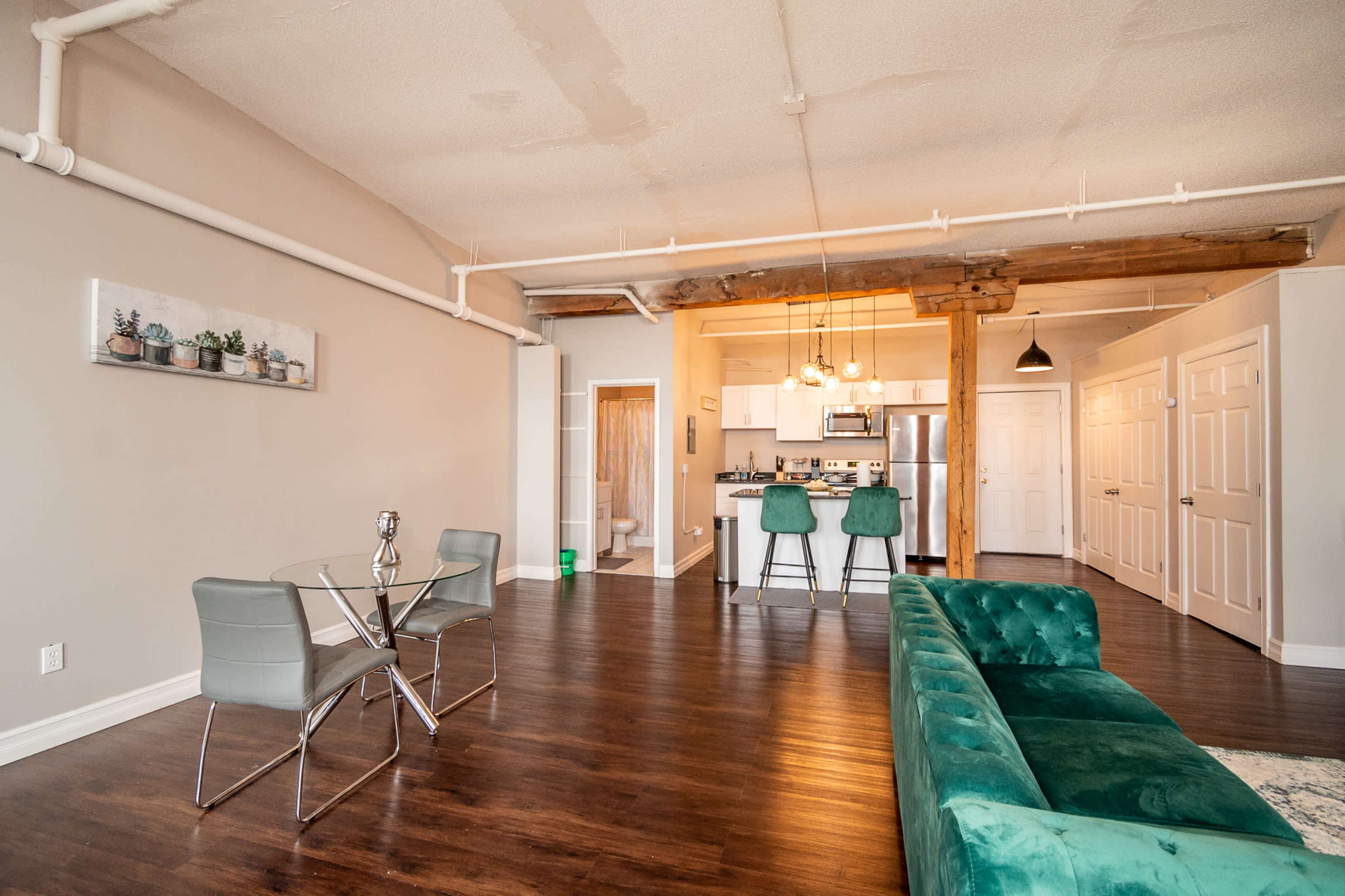 A modern living space featuring a green velvet sofa, a glass dining table with chairs, and an open kitchen area illuminated by pendant lights.