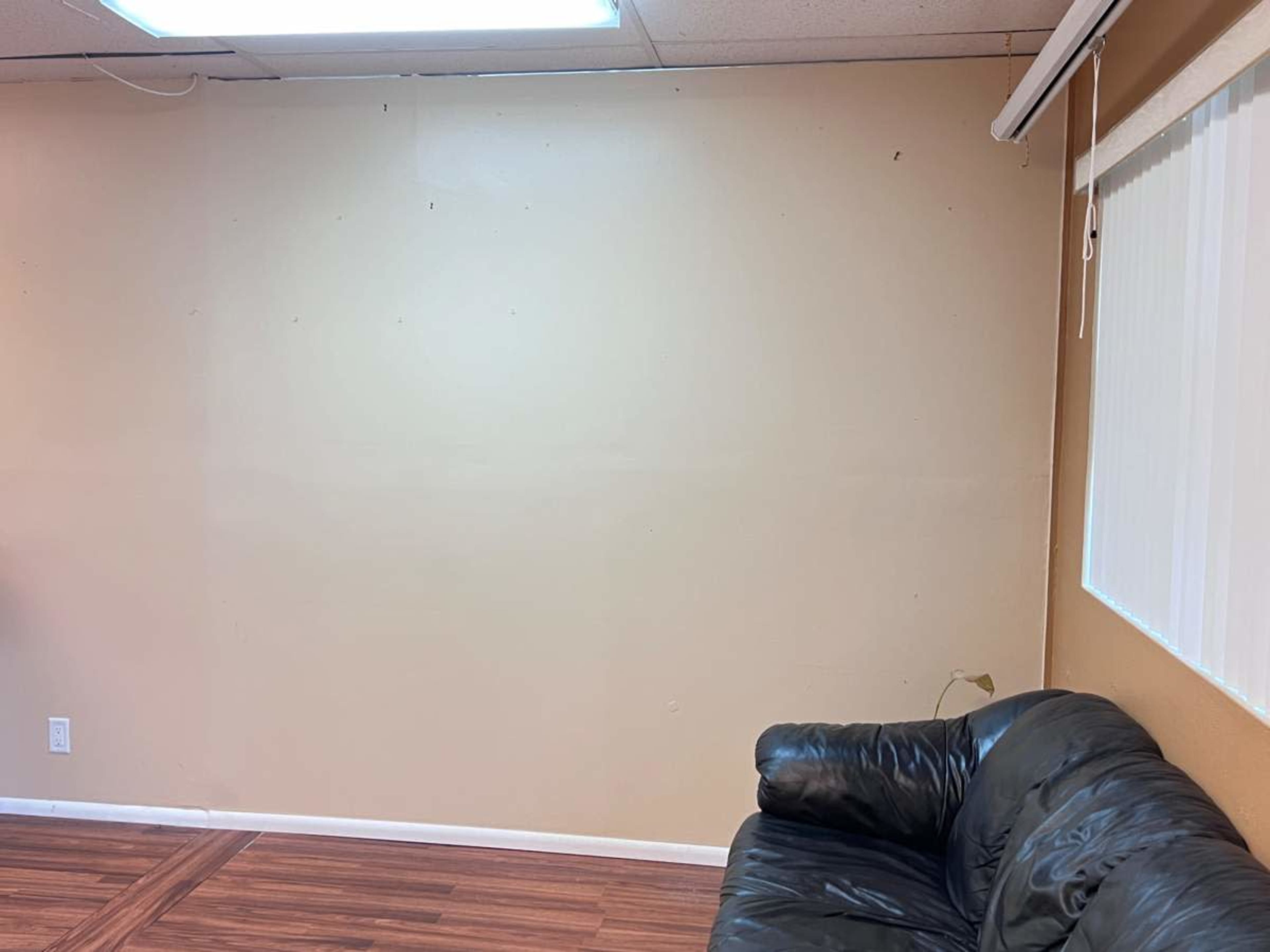 A plain beige wall is next to a black leather couch in a room with wooden flooring.