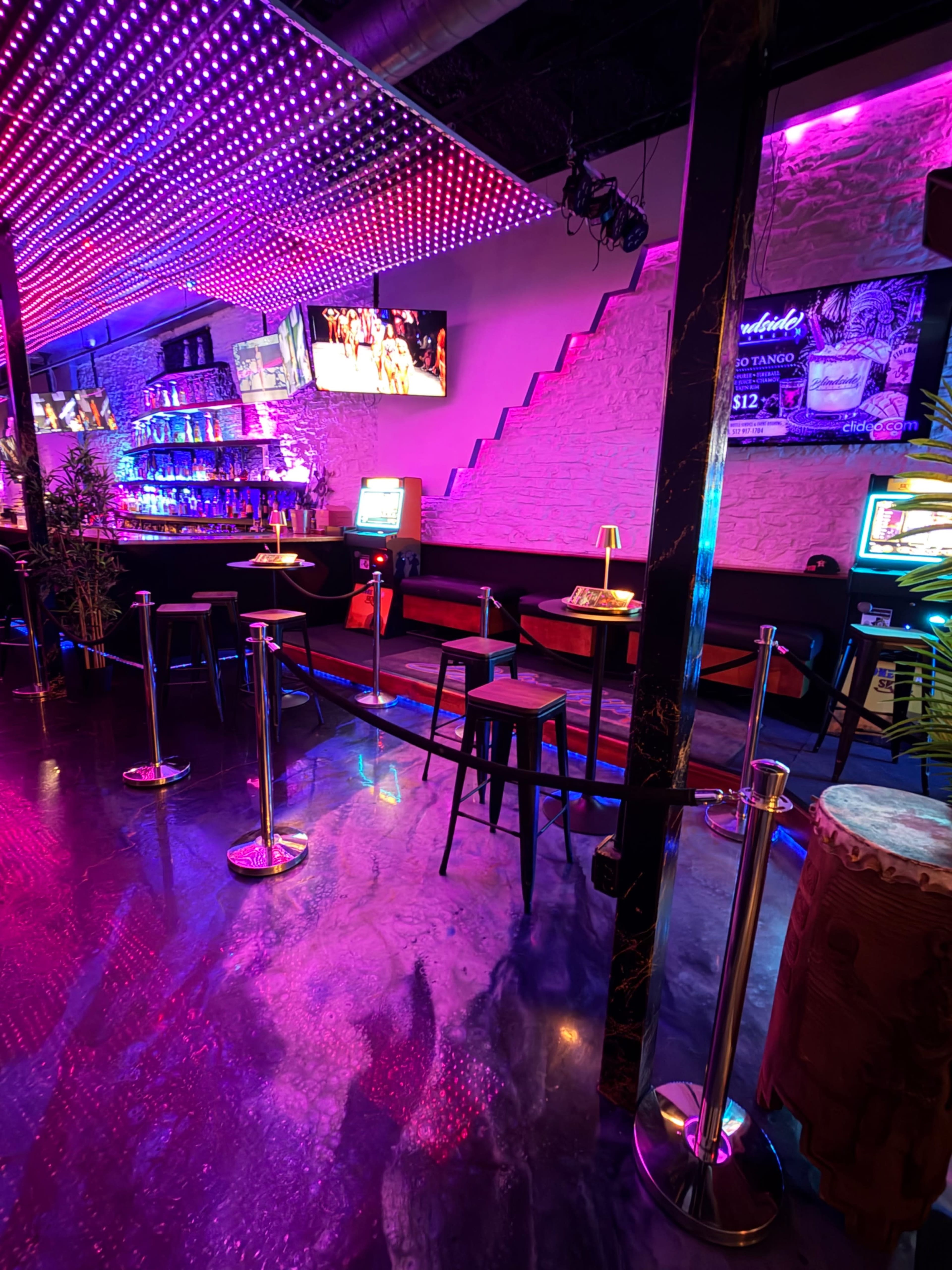 The image shows a lively bar scene with colorful LED lighting, a seated area along a wall, and a few bar stools facing a vibrant backlit counter.