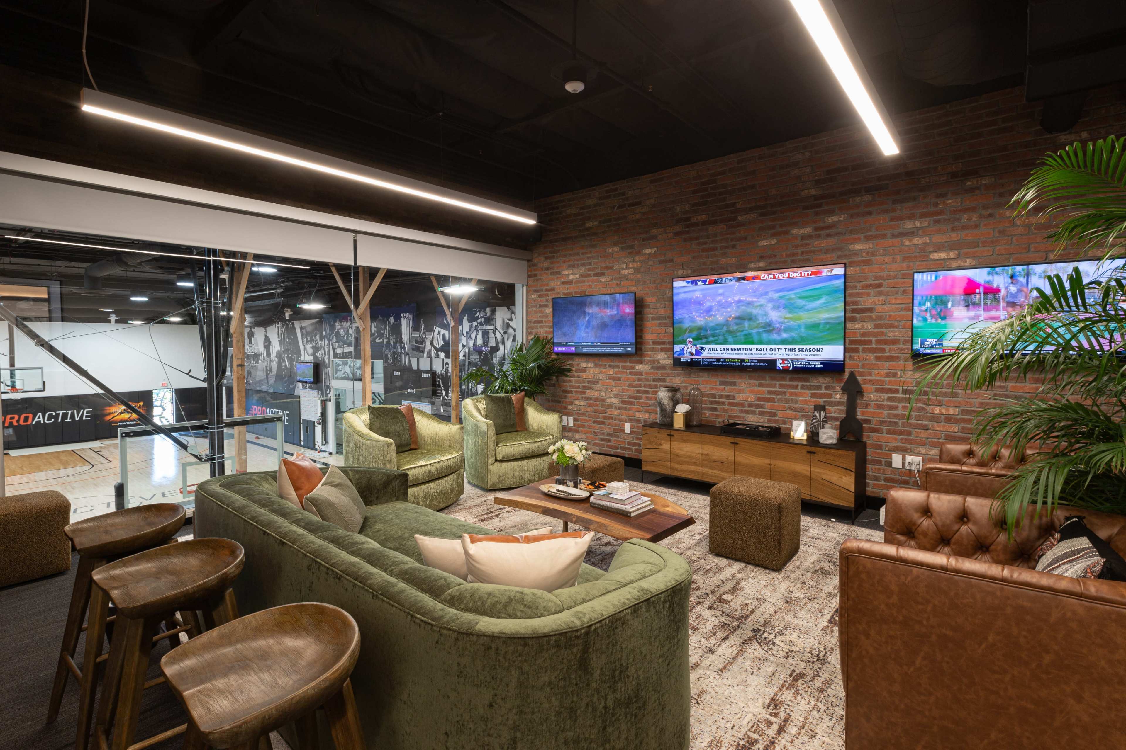 A modern lounge area features green sofas, a brown leather armchair, a wooden coffee table, and two televisions mounted on a brick wall, with a view of a sports court in the background.