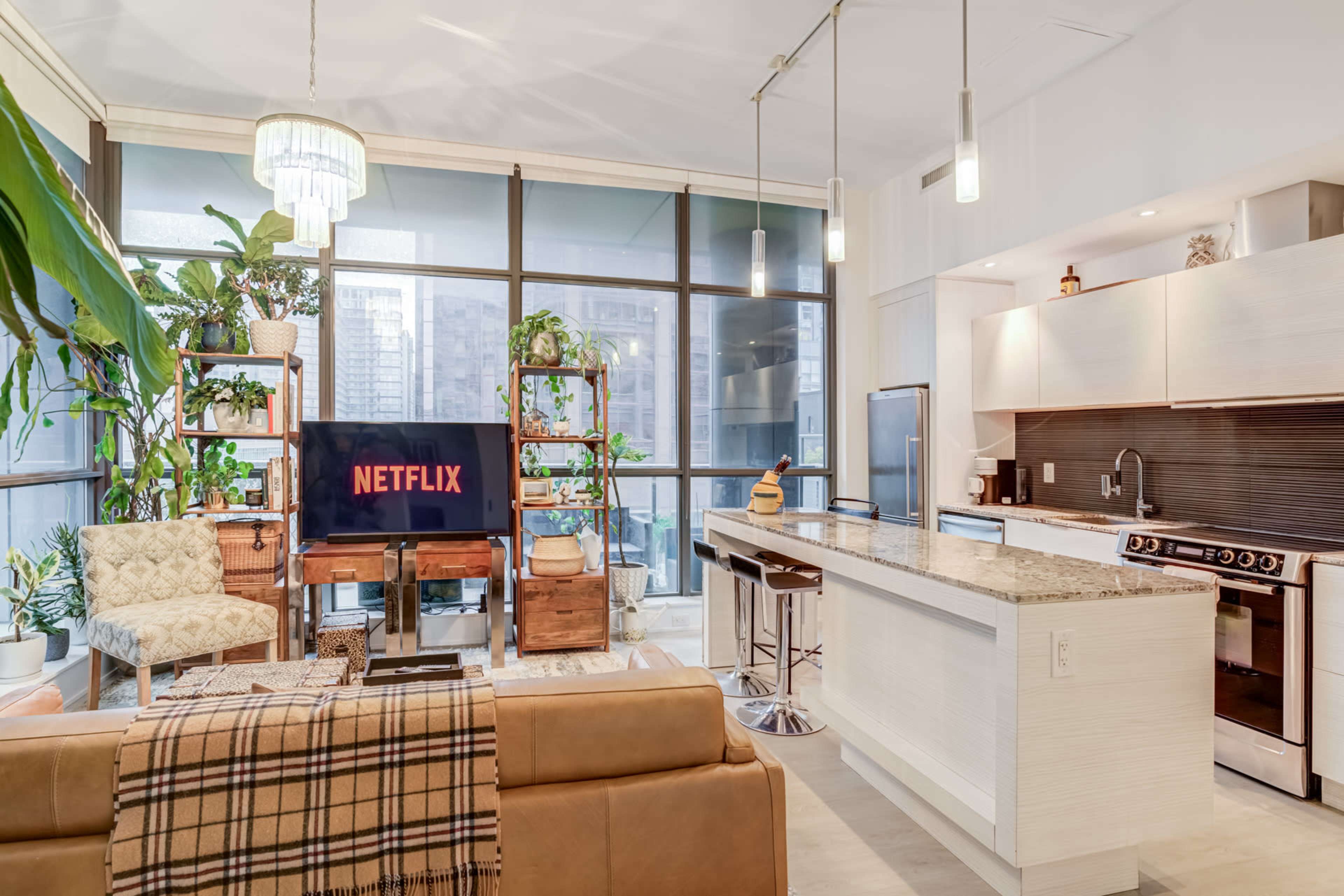 The image shows a modern living space with a kitchen, featuring a large television displaying the Netflix logo, surrounded by plants, a couch, and bar stools at the countertop.