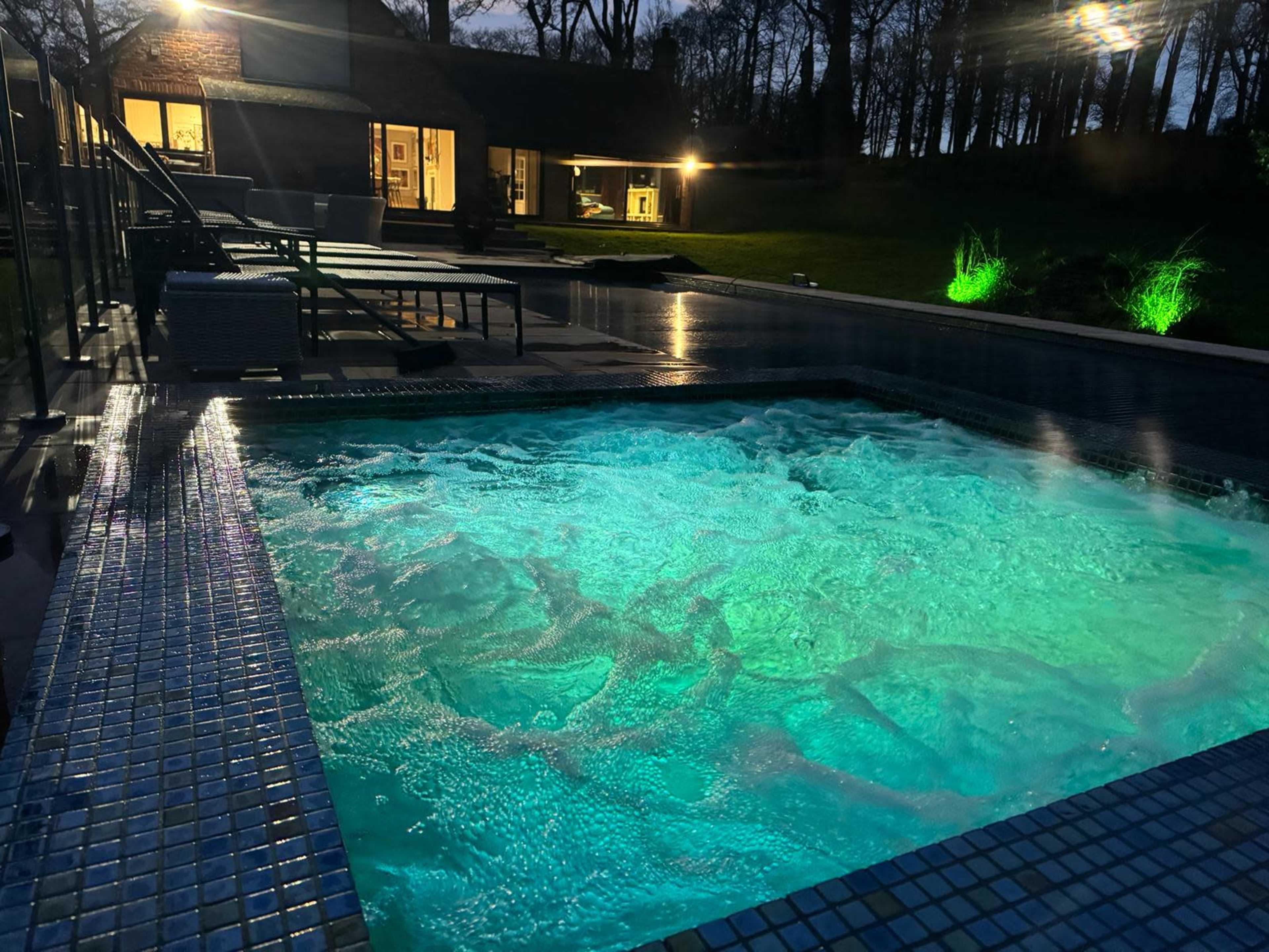 A hot tub with bubbling water is illuminated by green lighting, surrounded by a deck and trees at night.