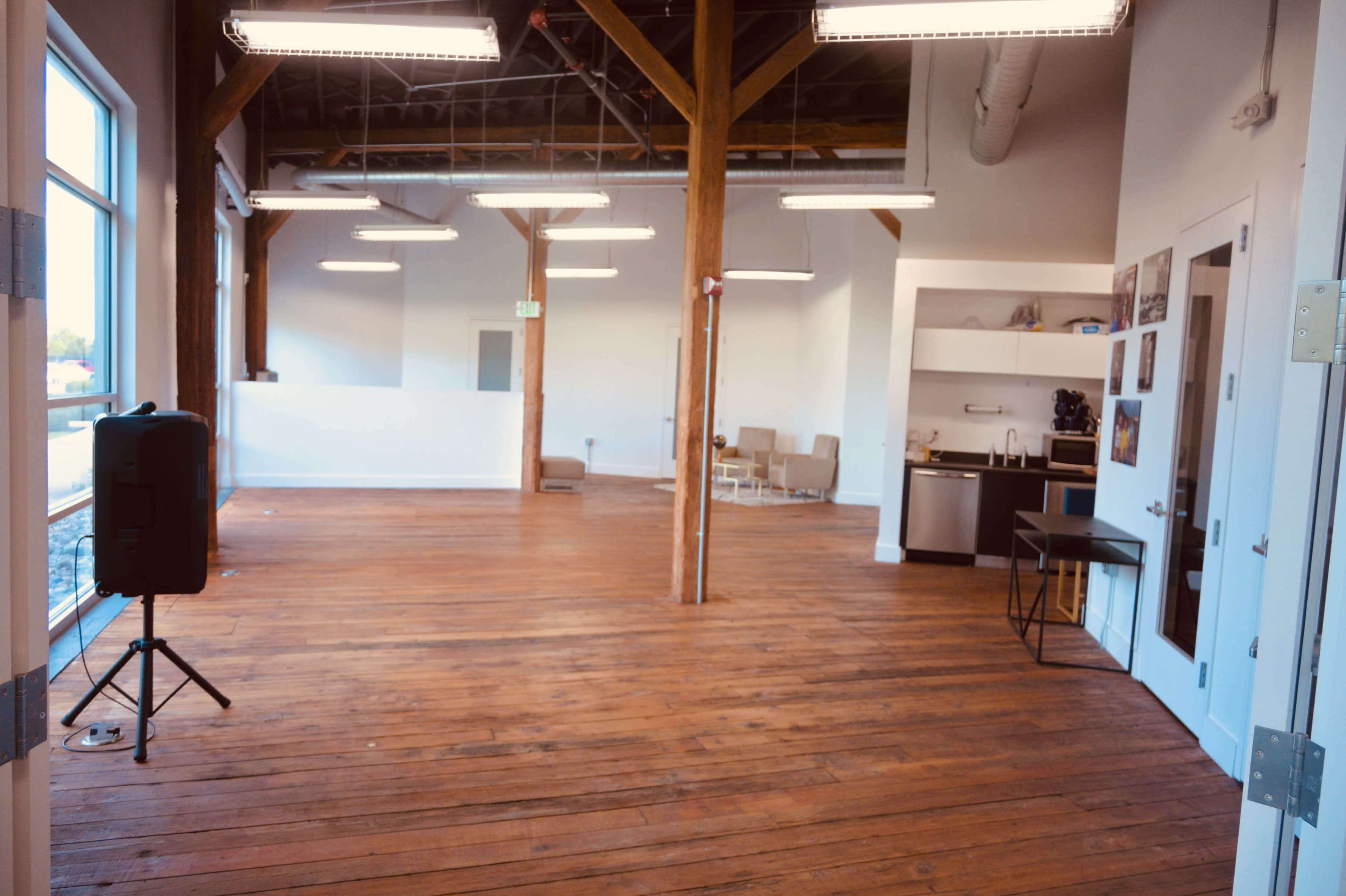 The image shows an open space with wooden flooring and exposed beams, featuring a kitchenette in the corner and some seating in the background.