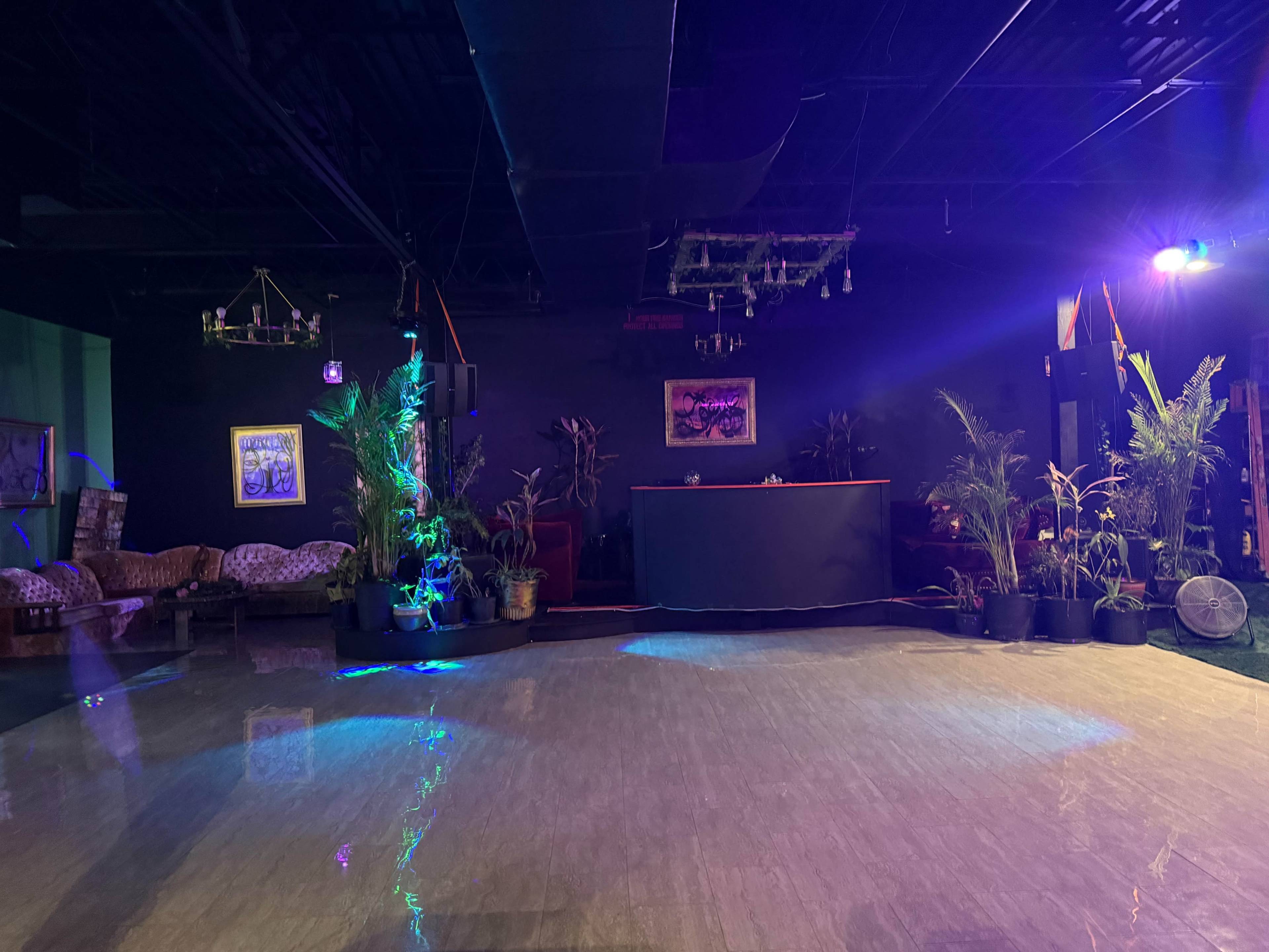 The image depicts an empty event space with a polished floor, seating area, and various potted plants under colorful lighting.
