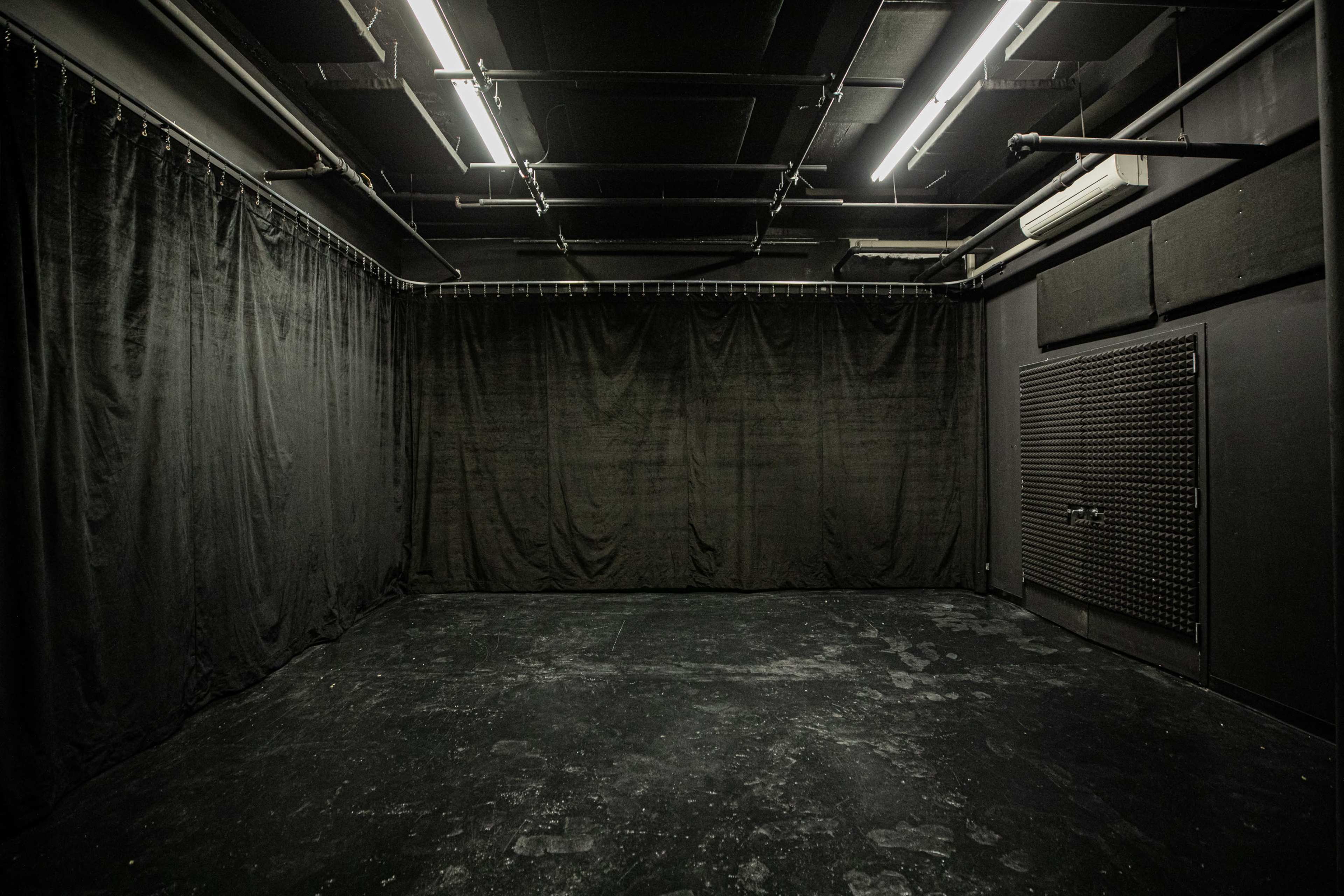 The image shows an empty room with black curtains lining the walls and a dark floor, illuminated by overhead fluorescent lights.