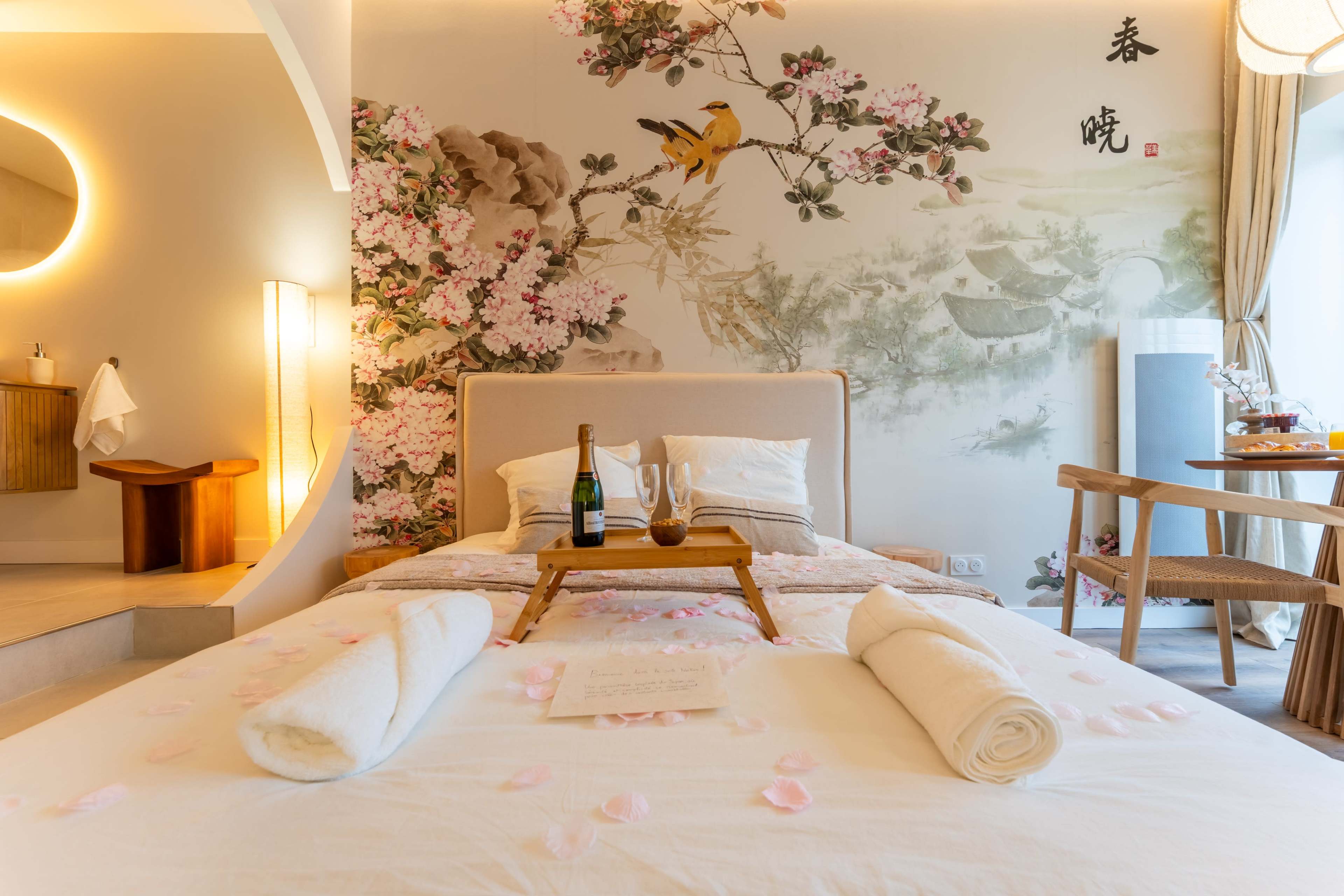 The image shows a beautifully arranged hotel room with a large bed adorned with rose petals, a decorative wall mural, and a small table with a bottle of champagne and glasses.