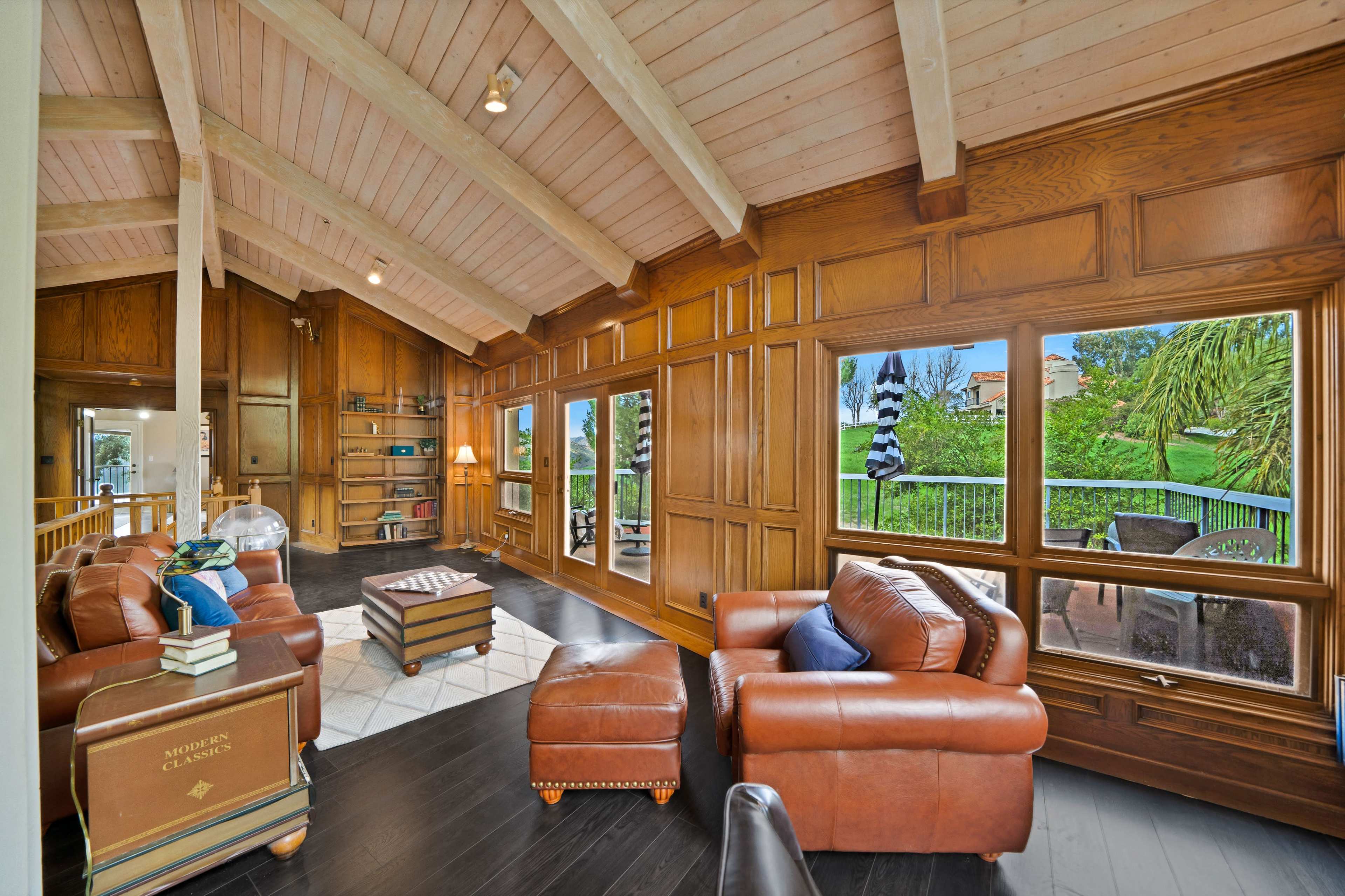 The interior of a spacious room features wooden wall paneling, large windows providing natural light, and leather seating arrangements.