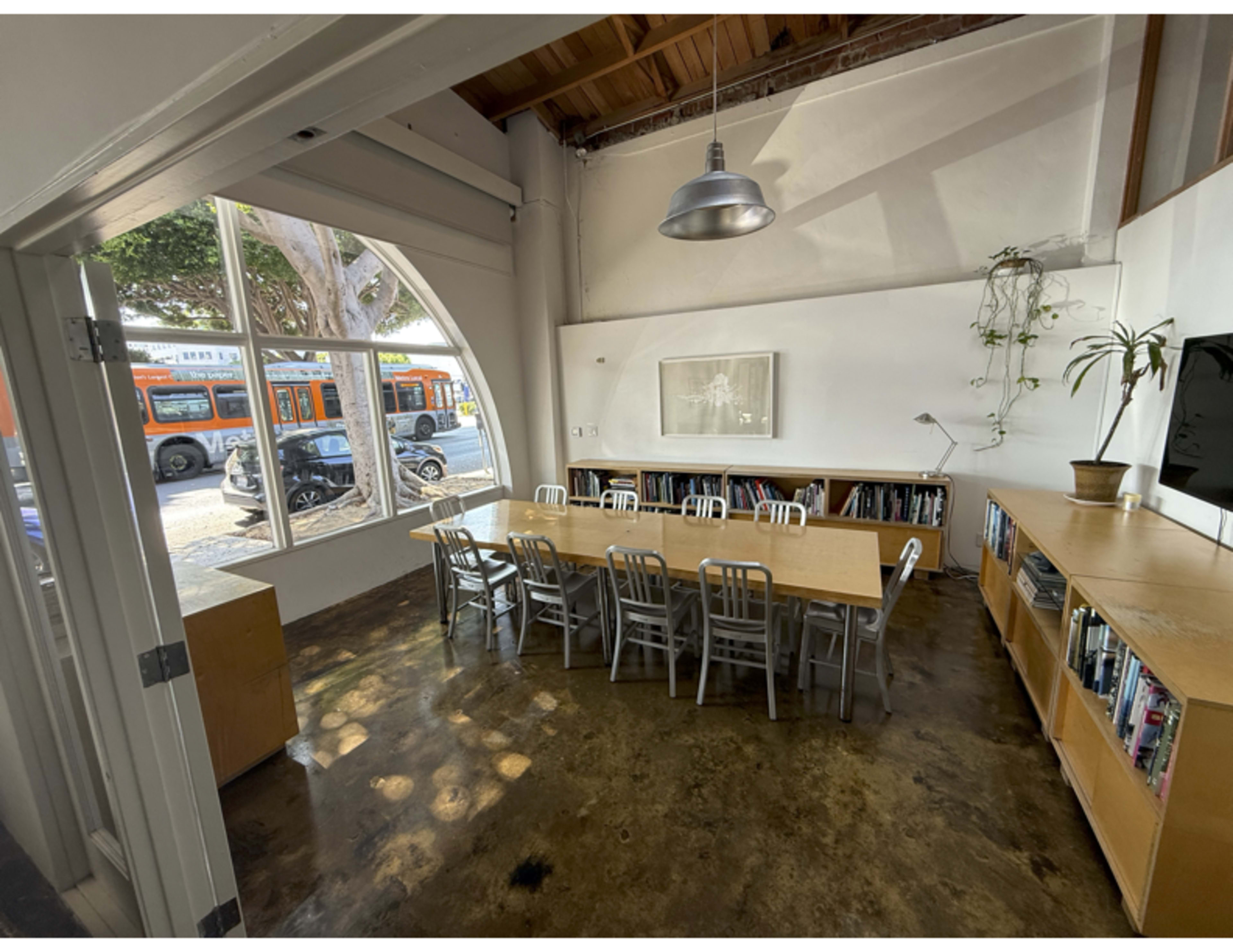 A spacious meeting room features a long wooden table surrounded by metal chairs, with large windows offering a view of the street outside.