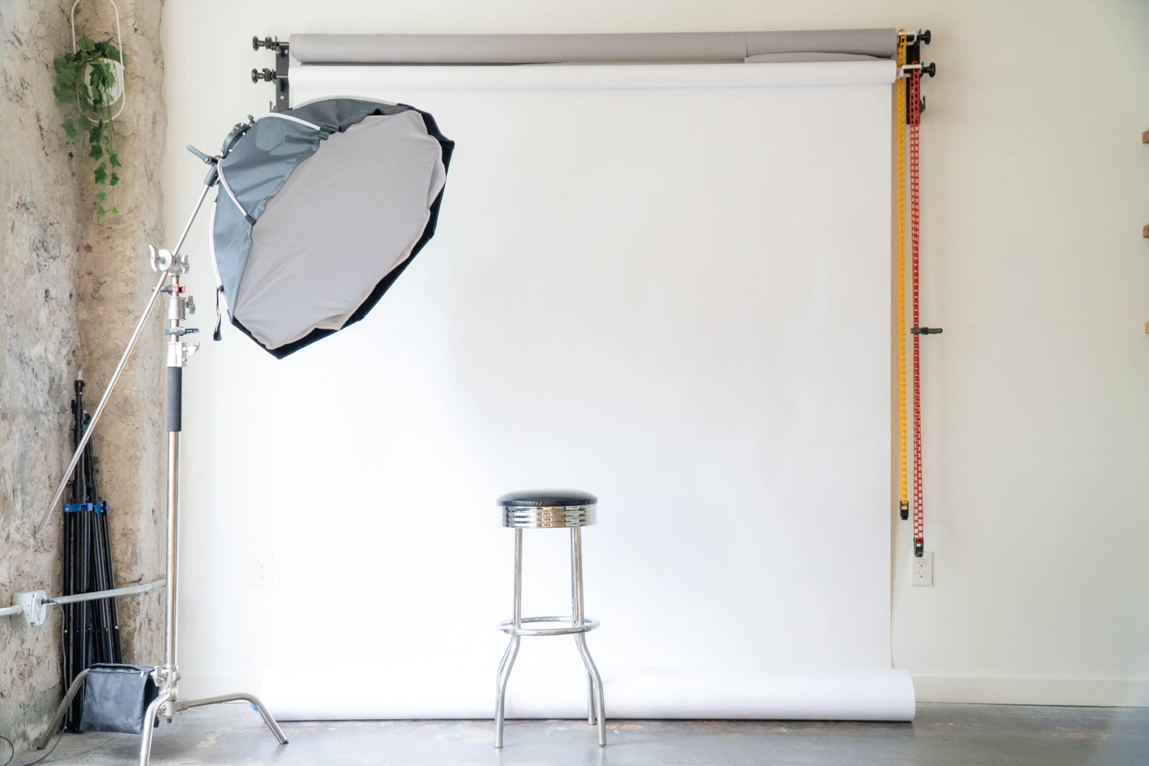 The image shows a photography studio setup featuring a white backdrop, a silver stool, and a light reflector on a stand.