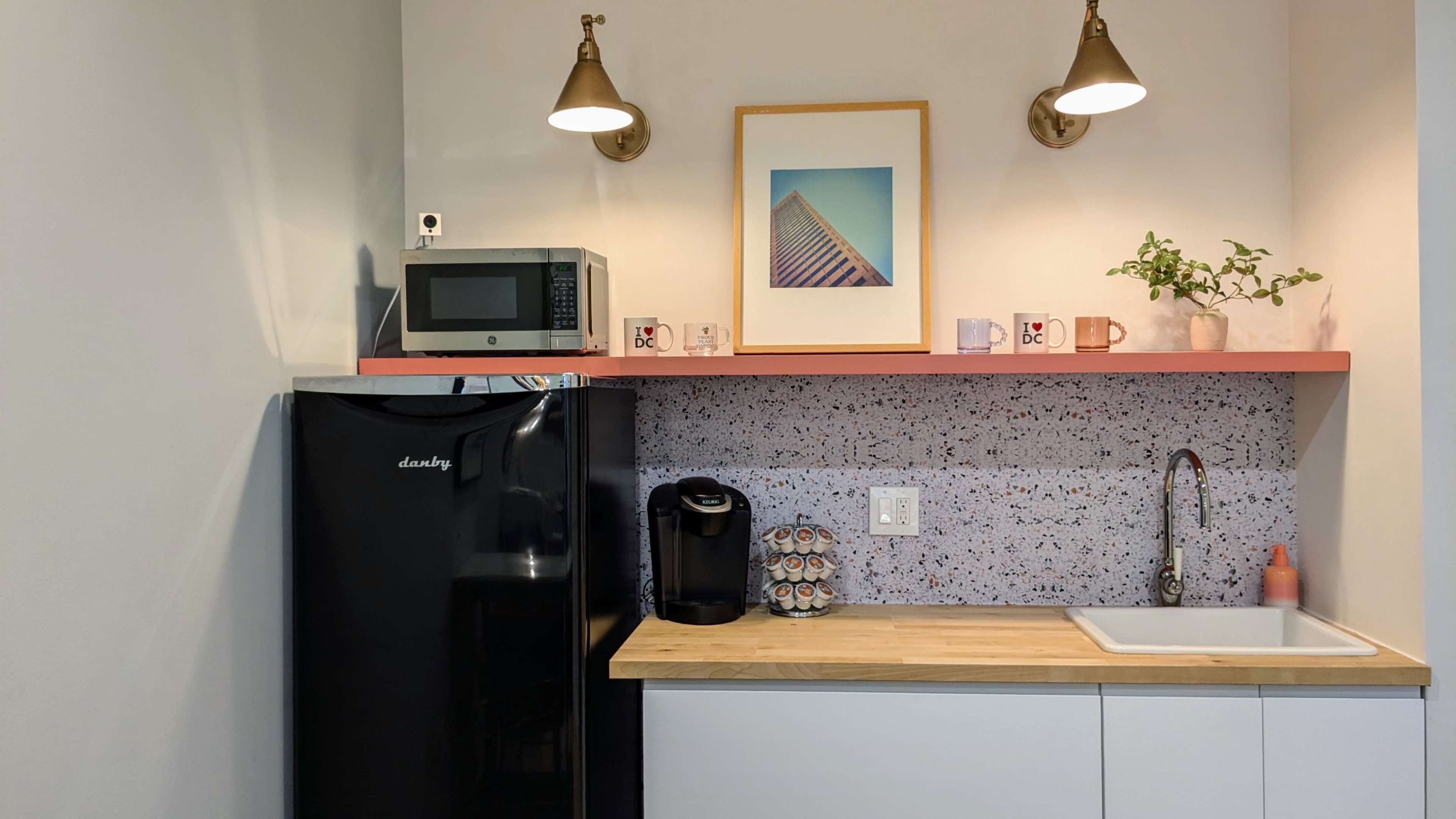 A modern kitchen corner features a black refrigerator, a countertop with a sink, a microwave, and a coffee maker, accented by a shelf displaying mugs and a framed artwork.