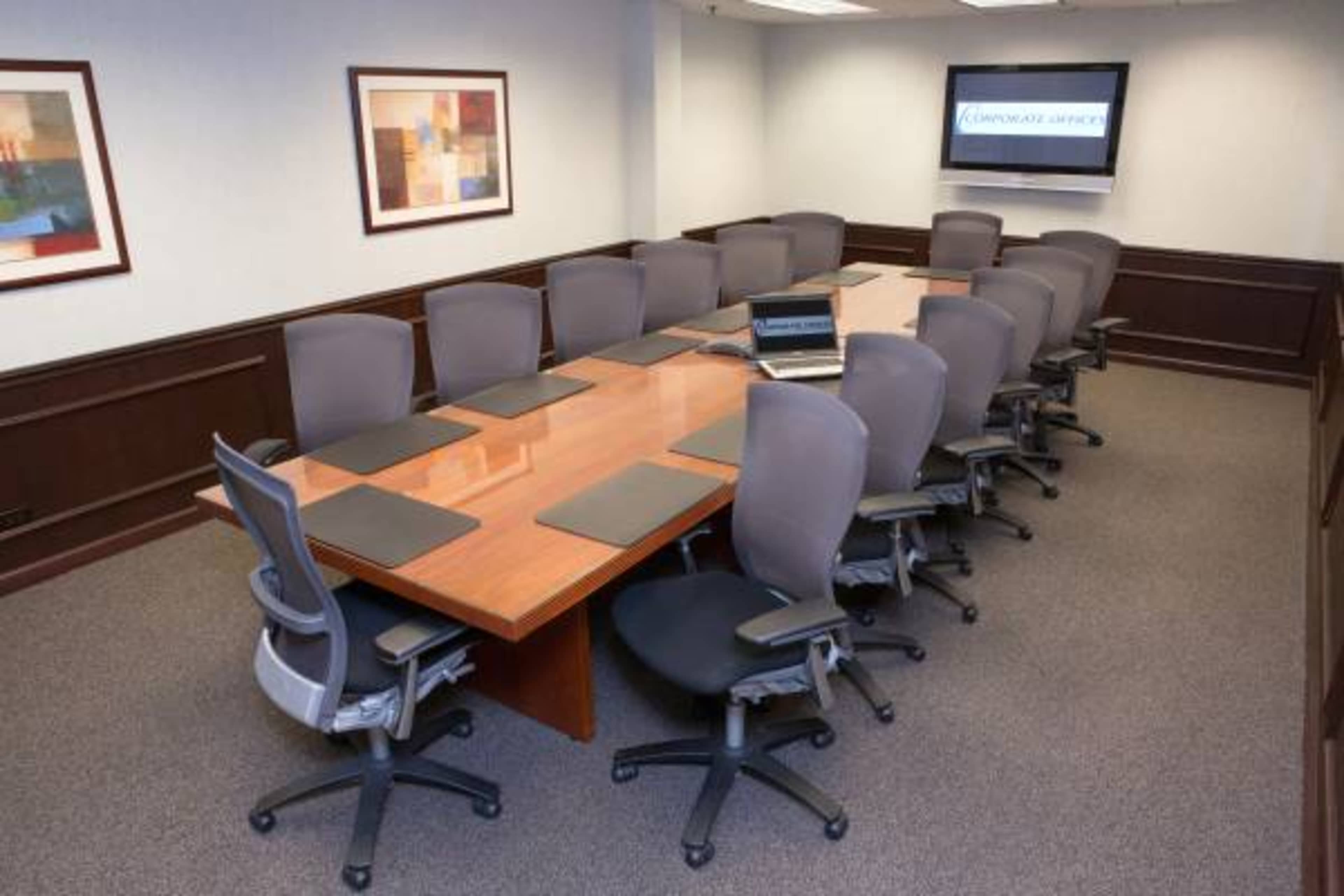 A large conference room features a long wooden table surrounded by ergonomic chairs, with an empty laptop placed on the table and a screen displaying a logo on the wall.