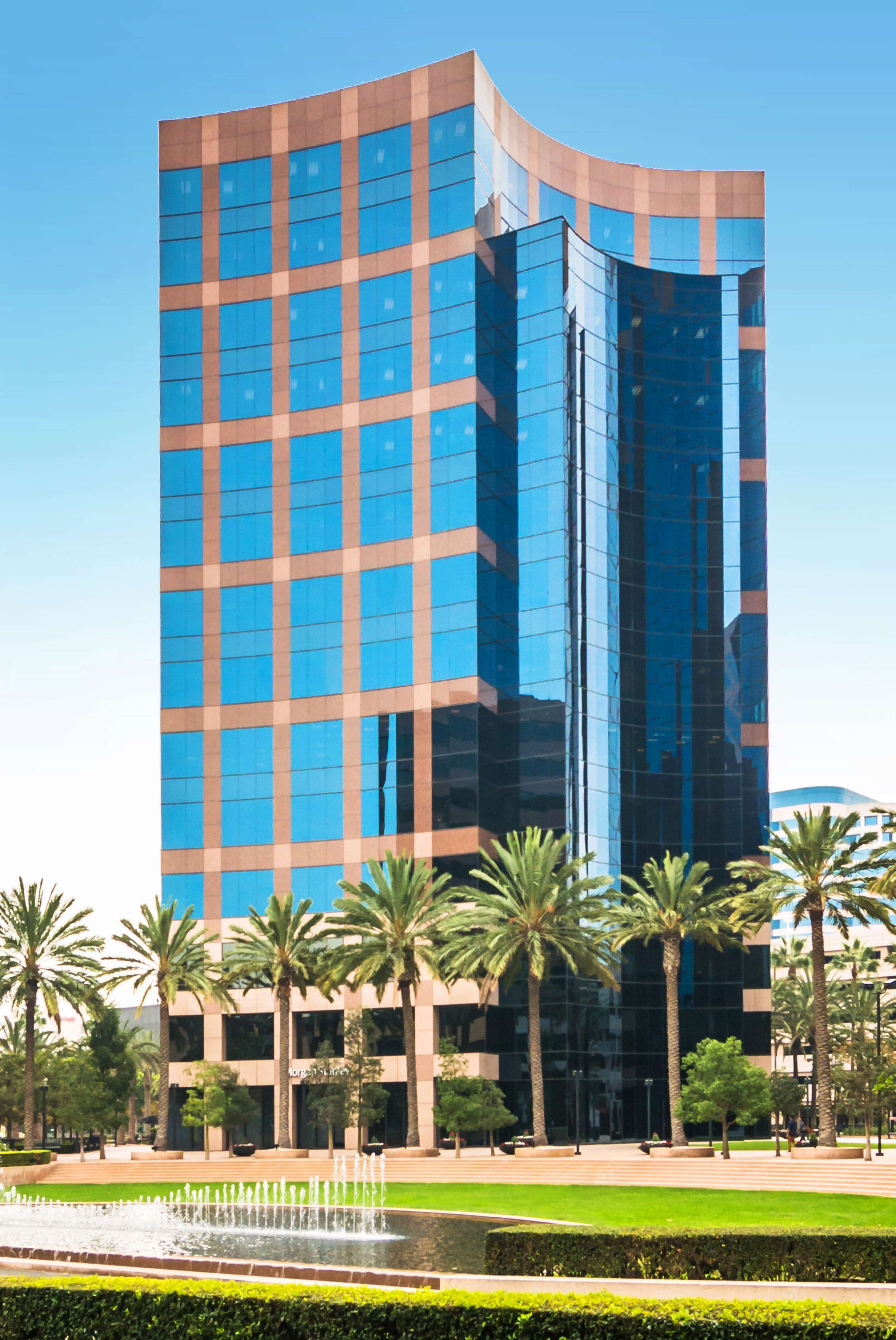 A modern glass-walled office building with a curved design is surrounded by palm trees and a landscaped plaza featuring water fountains.