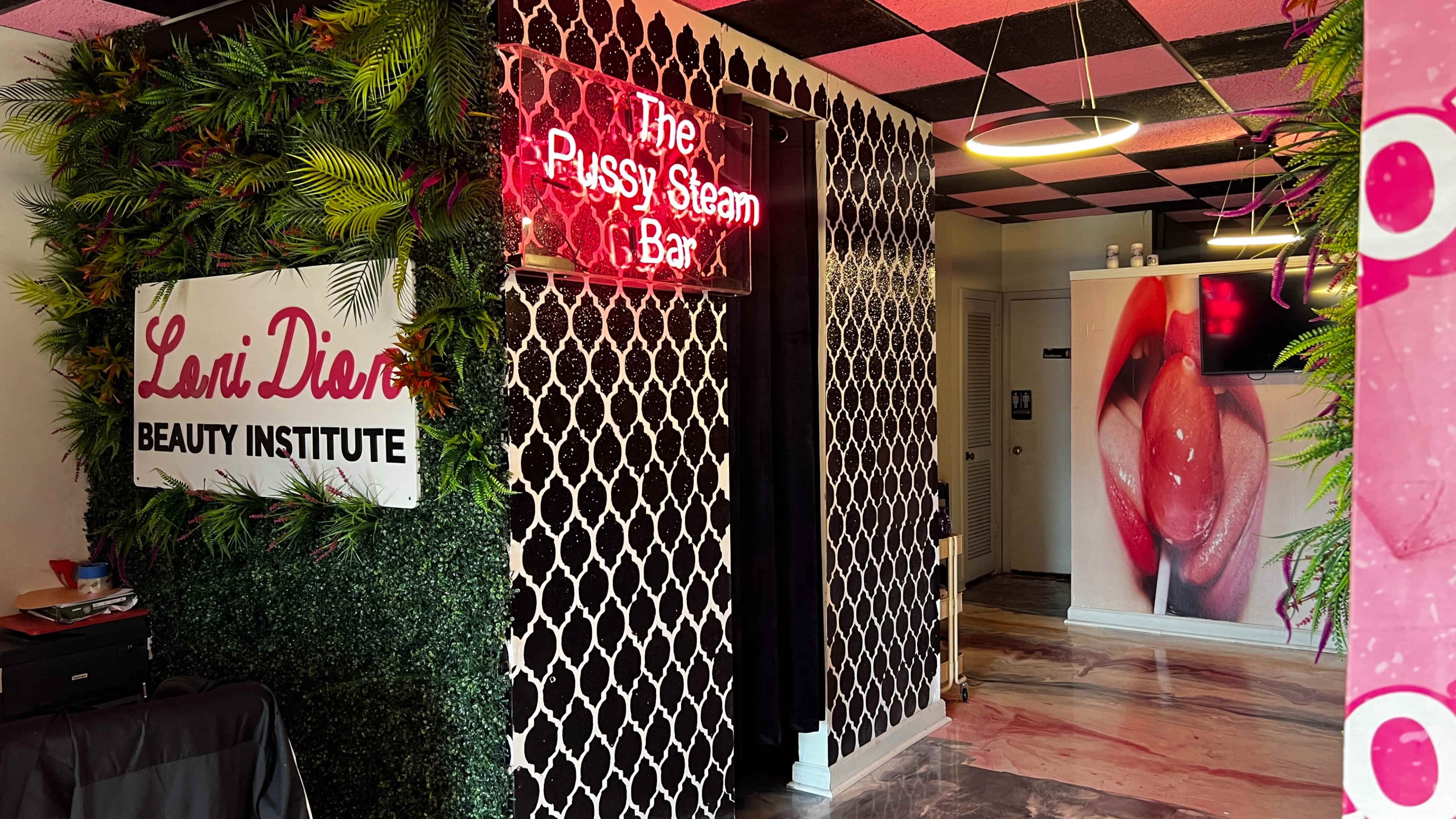 The entrance of a beauty institute featuring a neon sign for "The Pussy Steam Bar" amidst a decorated wall and lush greenery.