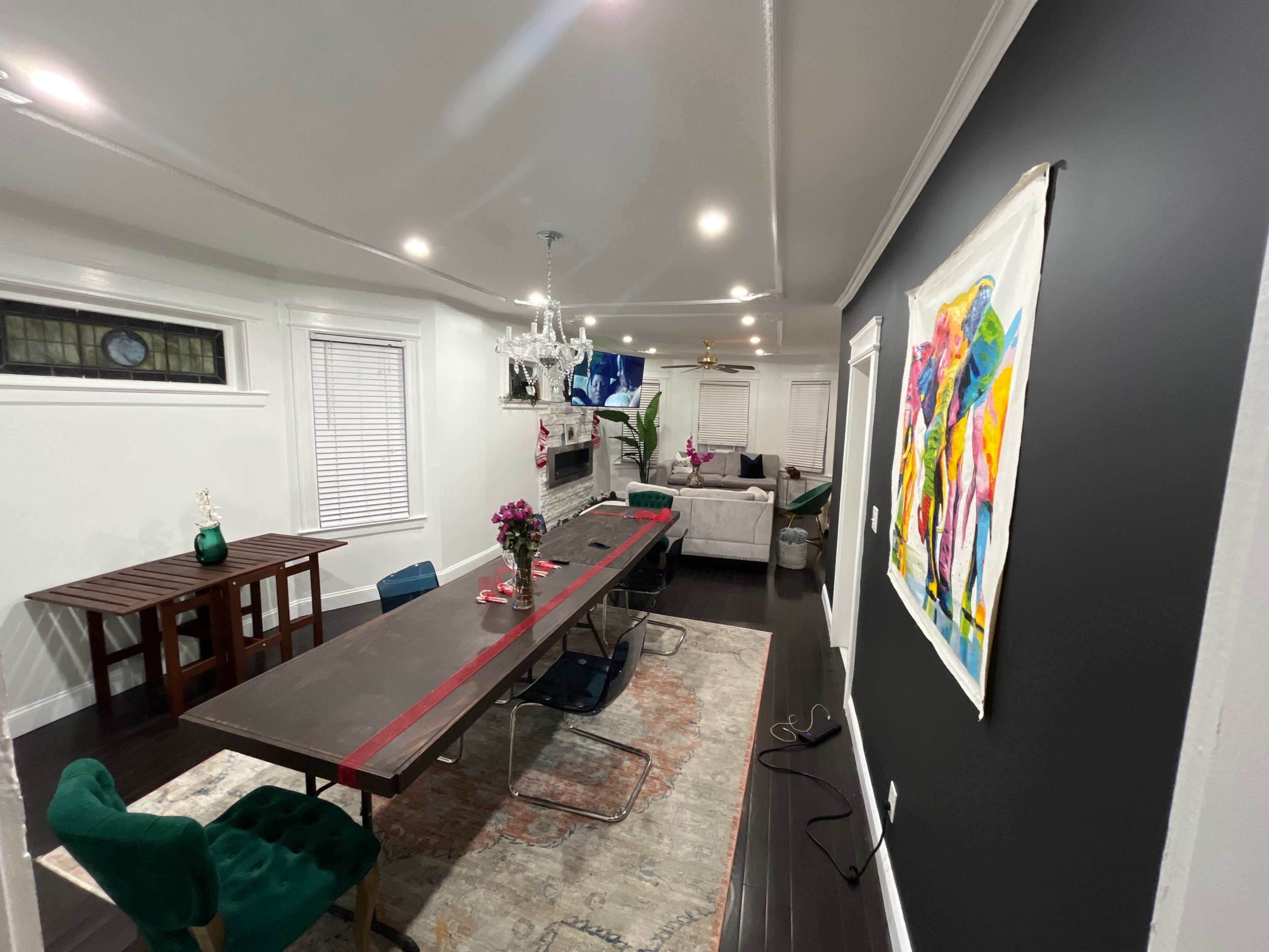 A modern dining area with a long table, colorful chairs, a chandelier, and a framed artwork on a dark wall.