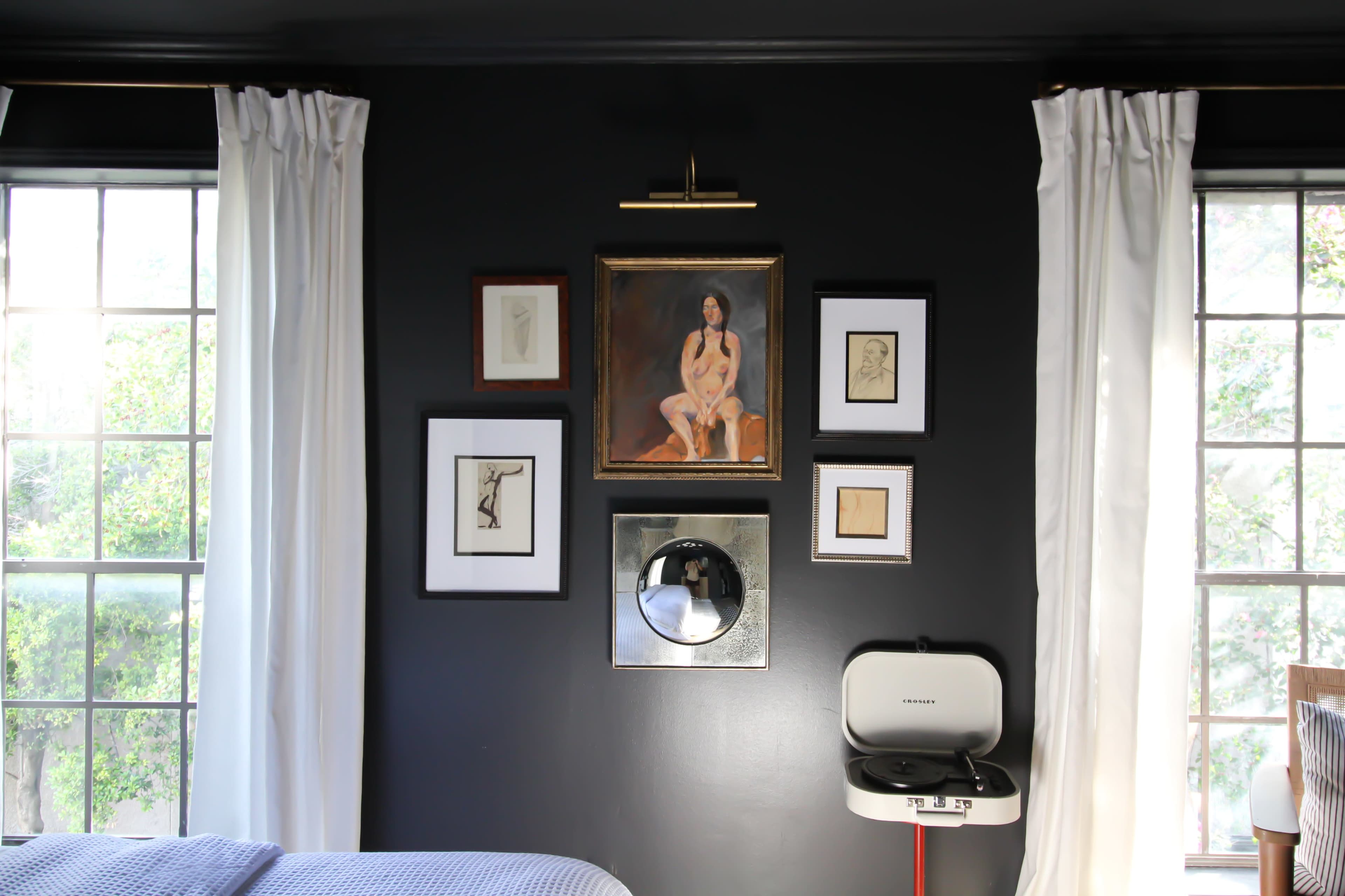 A dark room with two windows flanked by white curtains, featuring a gallery wall of framed artworks above a vintage-style mirror and a white bedside table.
