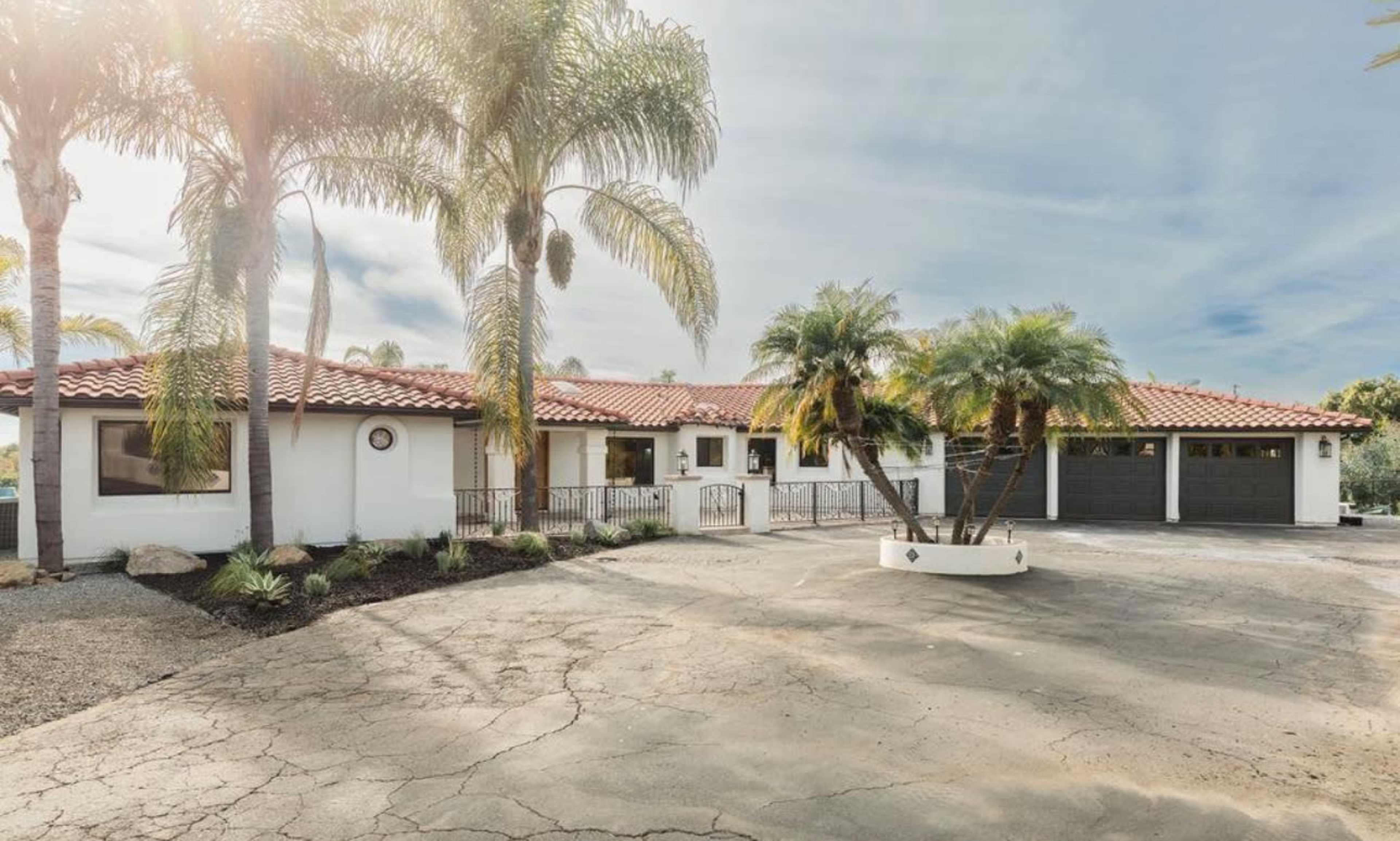 The image shows a single-story, Mediterranean-style house with palm trees in the front yard and a large, cracked driveway.