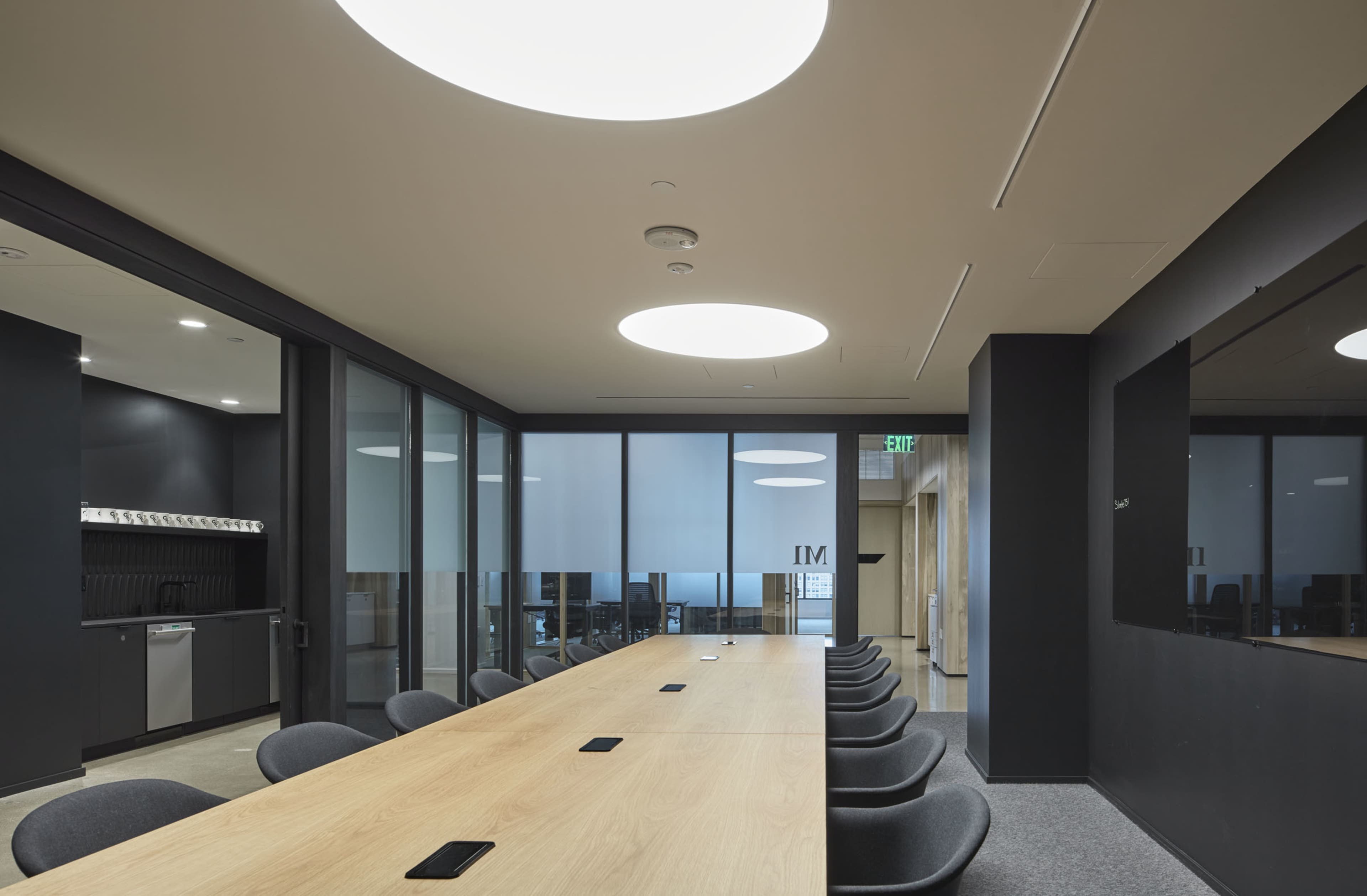 A modern conference room features a long wooden table surrounded by gray chairs, with large windows and minimalistic lighting.