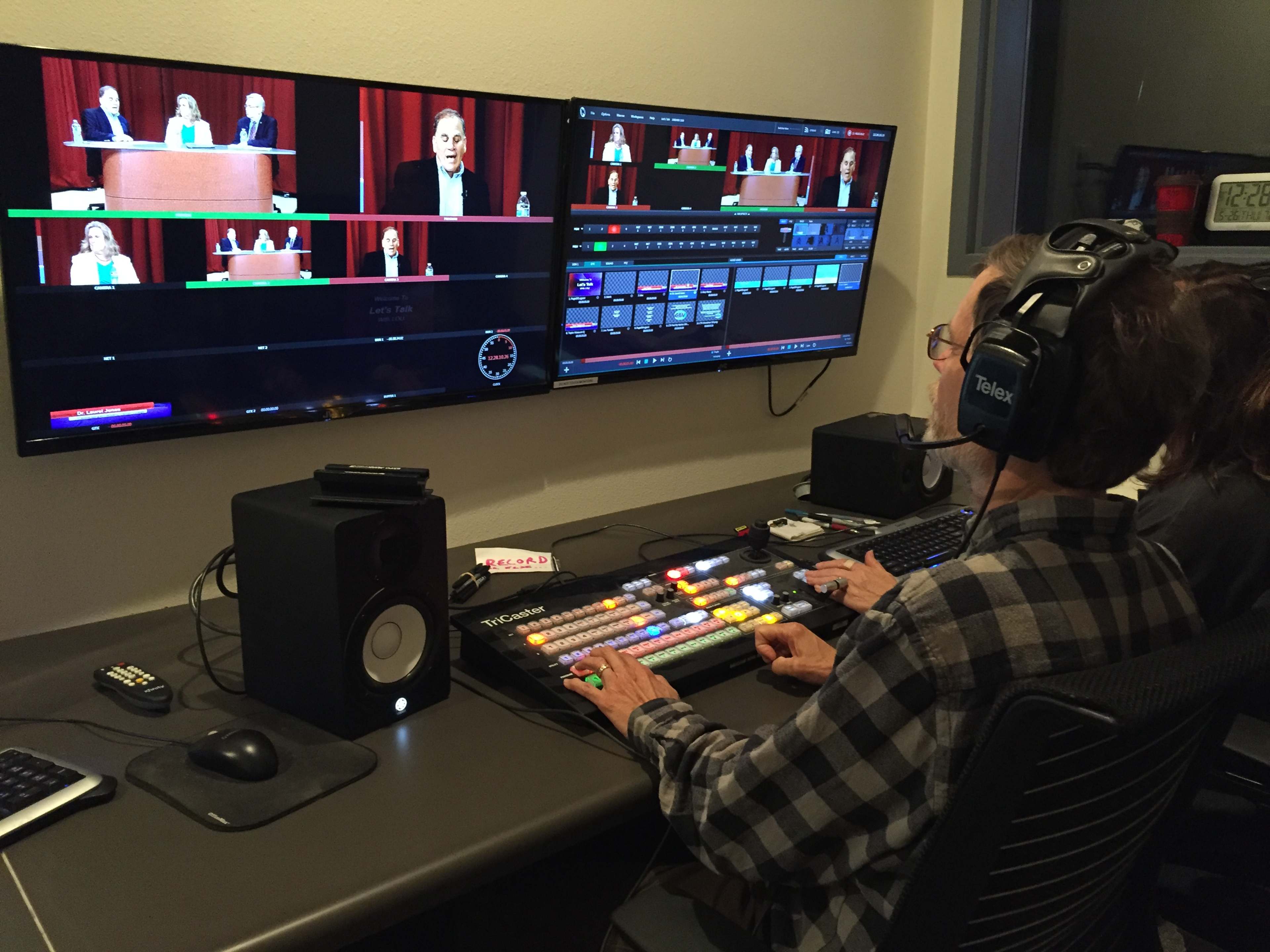 A person operates a video control panel while monitoring multiple screens displaying a live event with speakers at a podium.