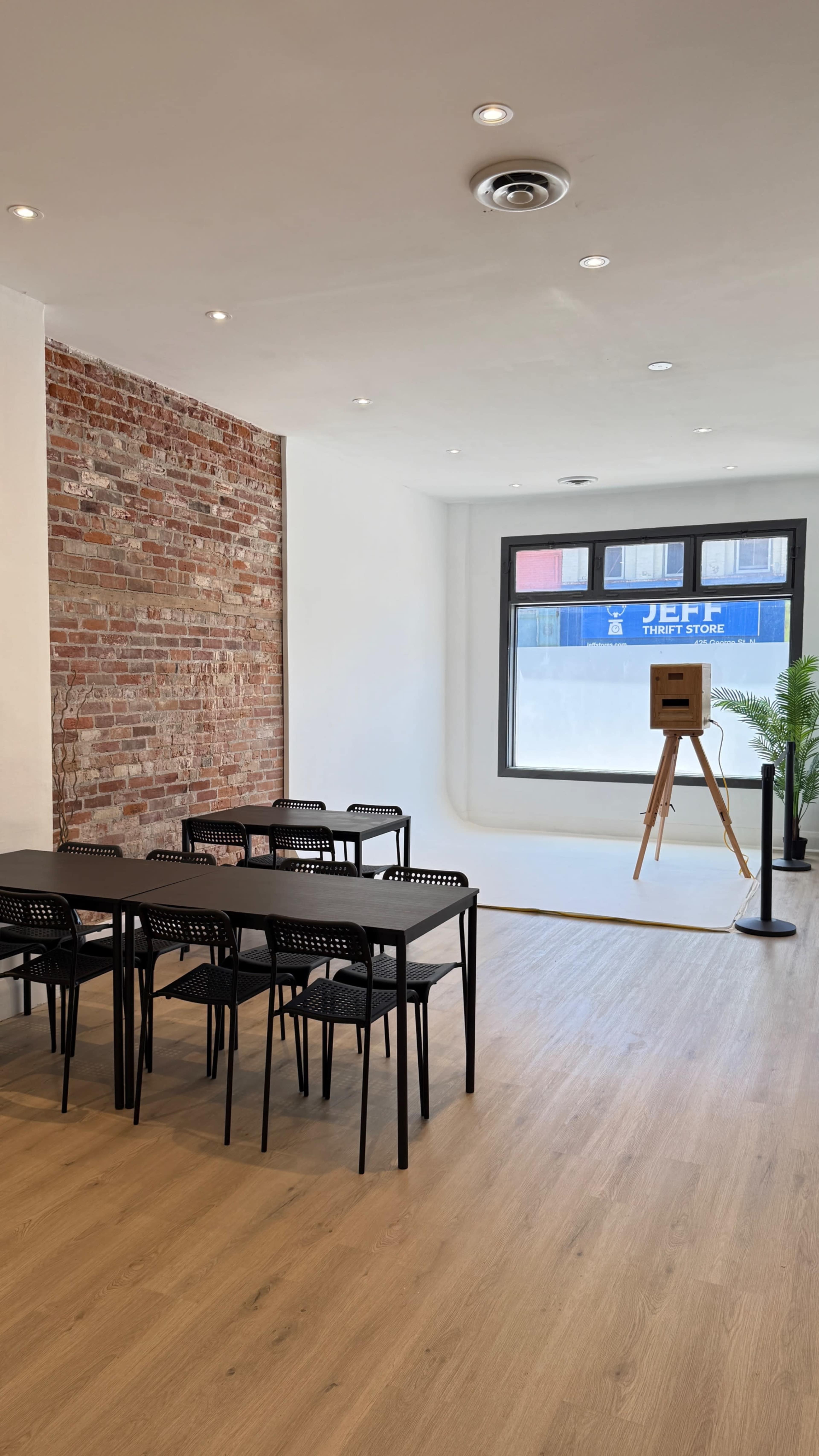 The image shows a modern, minimalist room with wooden flooring, featuring several black tables and chairs, a brick wall, and a large window displaying an easel.