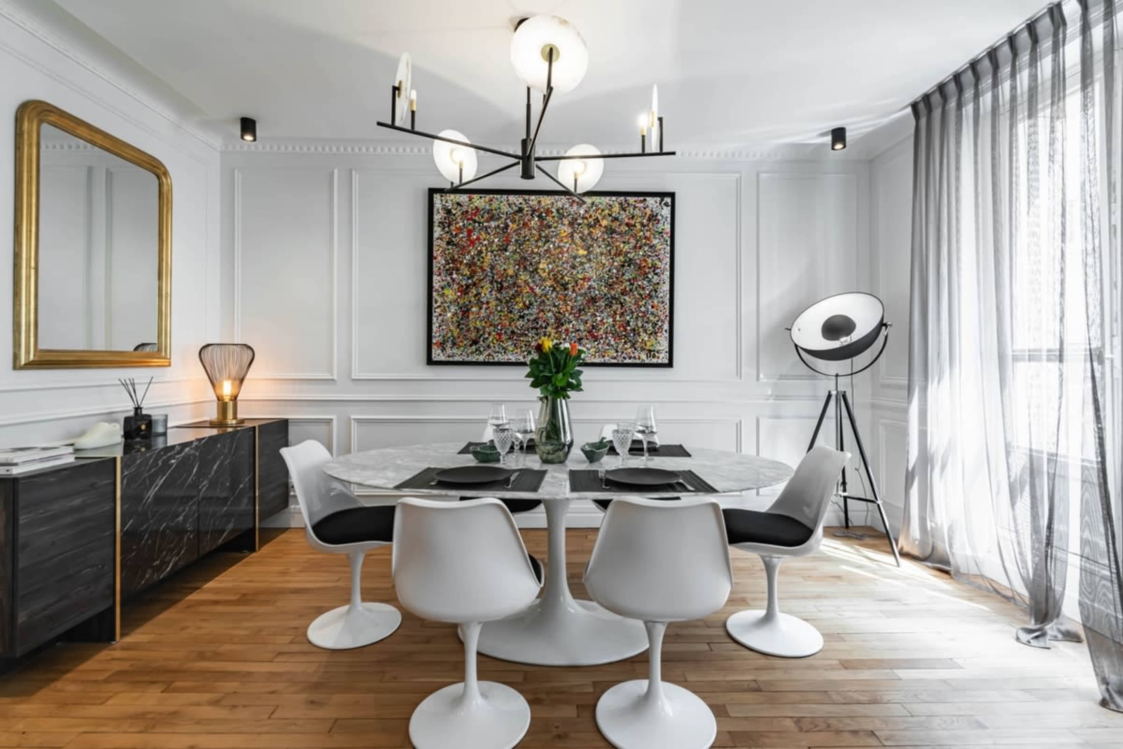 A modern dining room featuring a round marble dining table surrounded by white chairs, with a colorful abstract painting on the wall and a large window with sheer curtains.