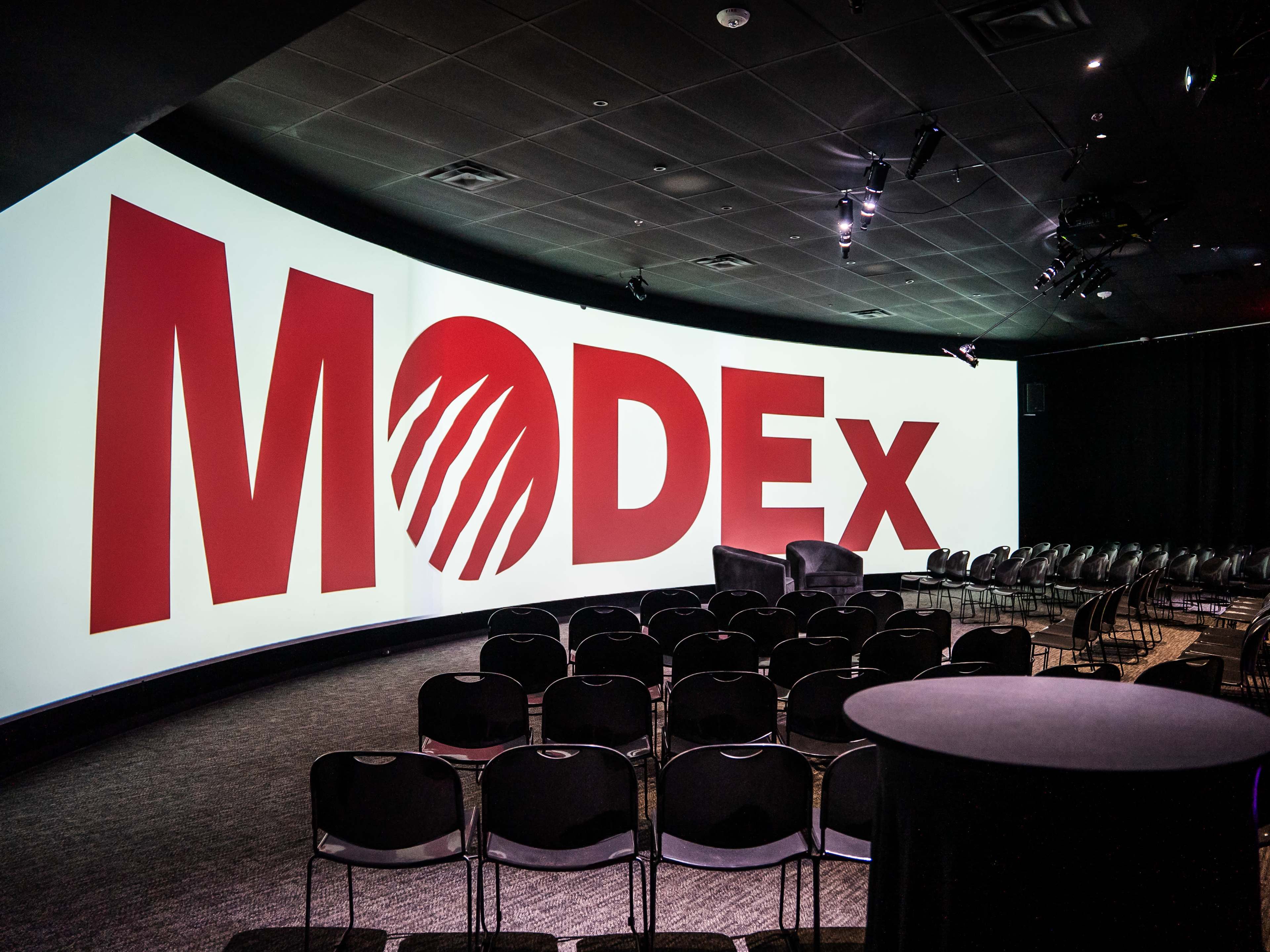 The image shows a conference room with a large curved screen displaying the word "MODEX" in bold red letters, surrounded by empty black chairs arranged for an event.