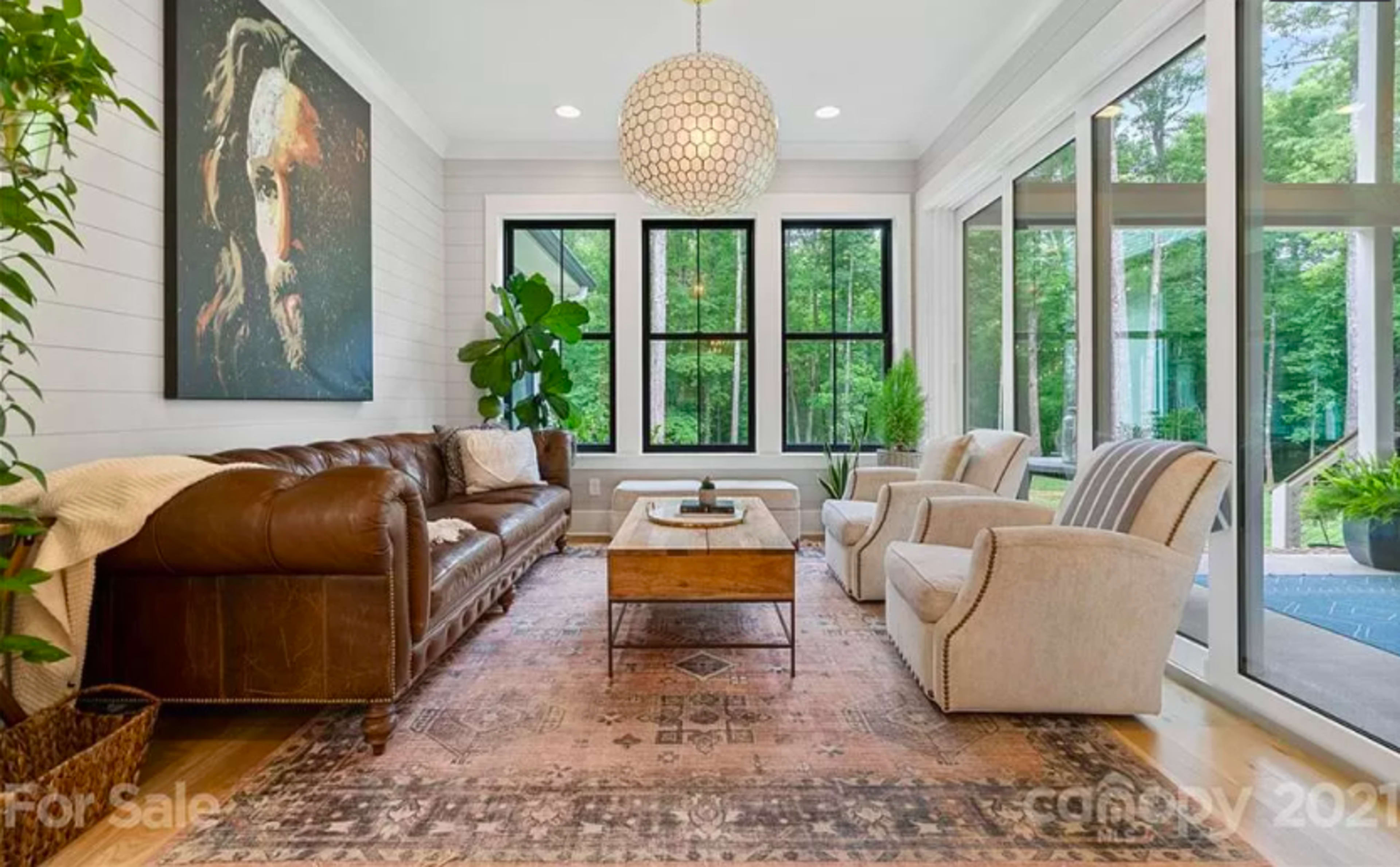 The living room features a brown leather sofa, two light-colored armchairs, a wooden coffee table, and large windows with outdoor greenery visible.
