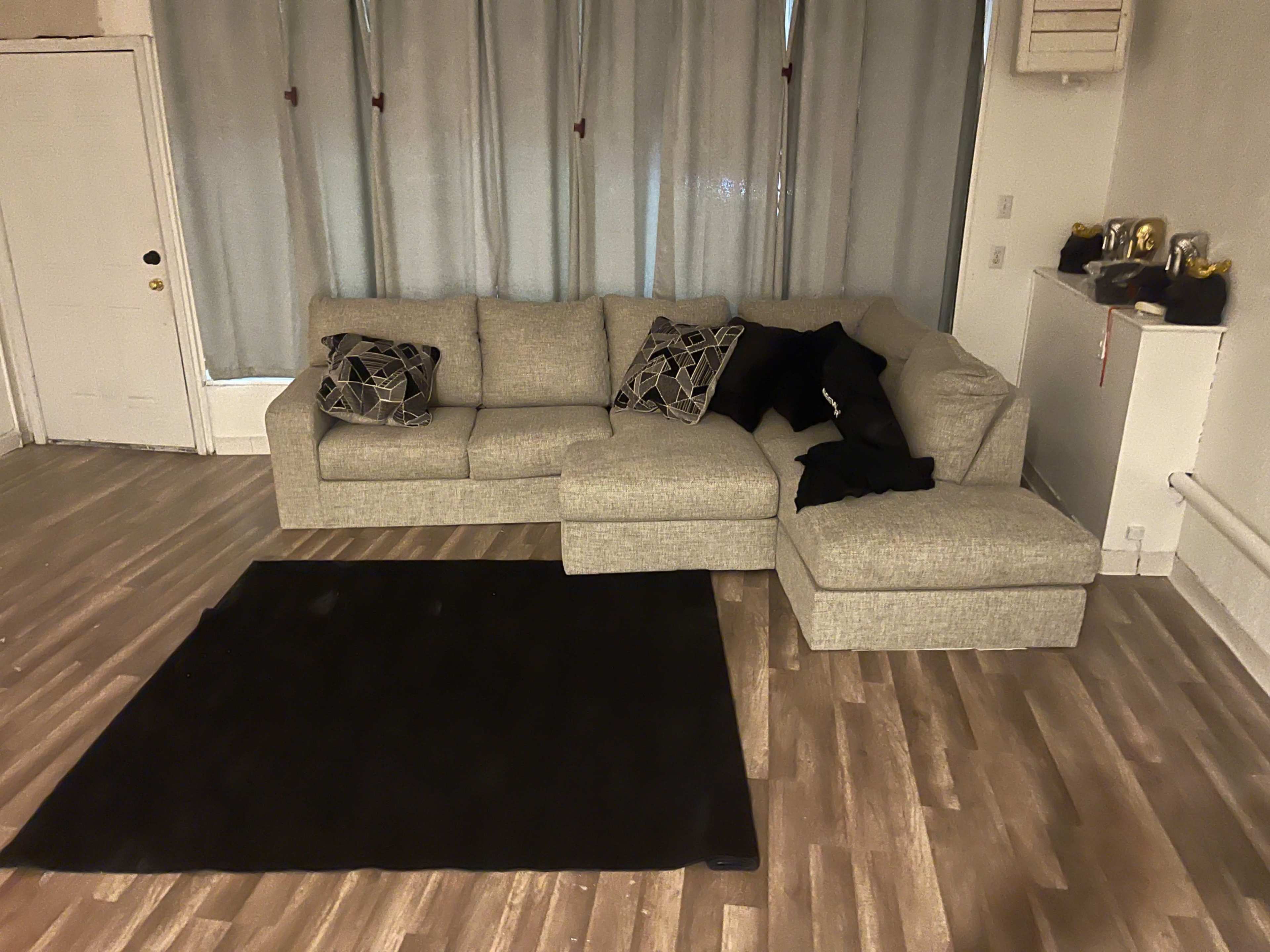 A sectional sofa with black and gray decorative pillows, positioned on a wooden floor in a room with light-colored walls and sheer curtains.