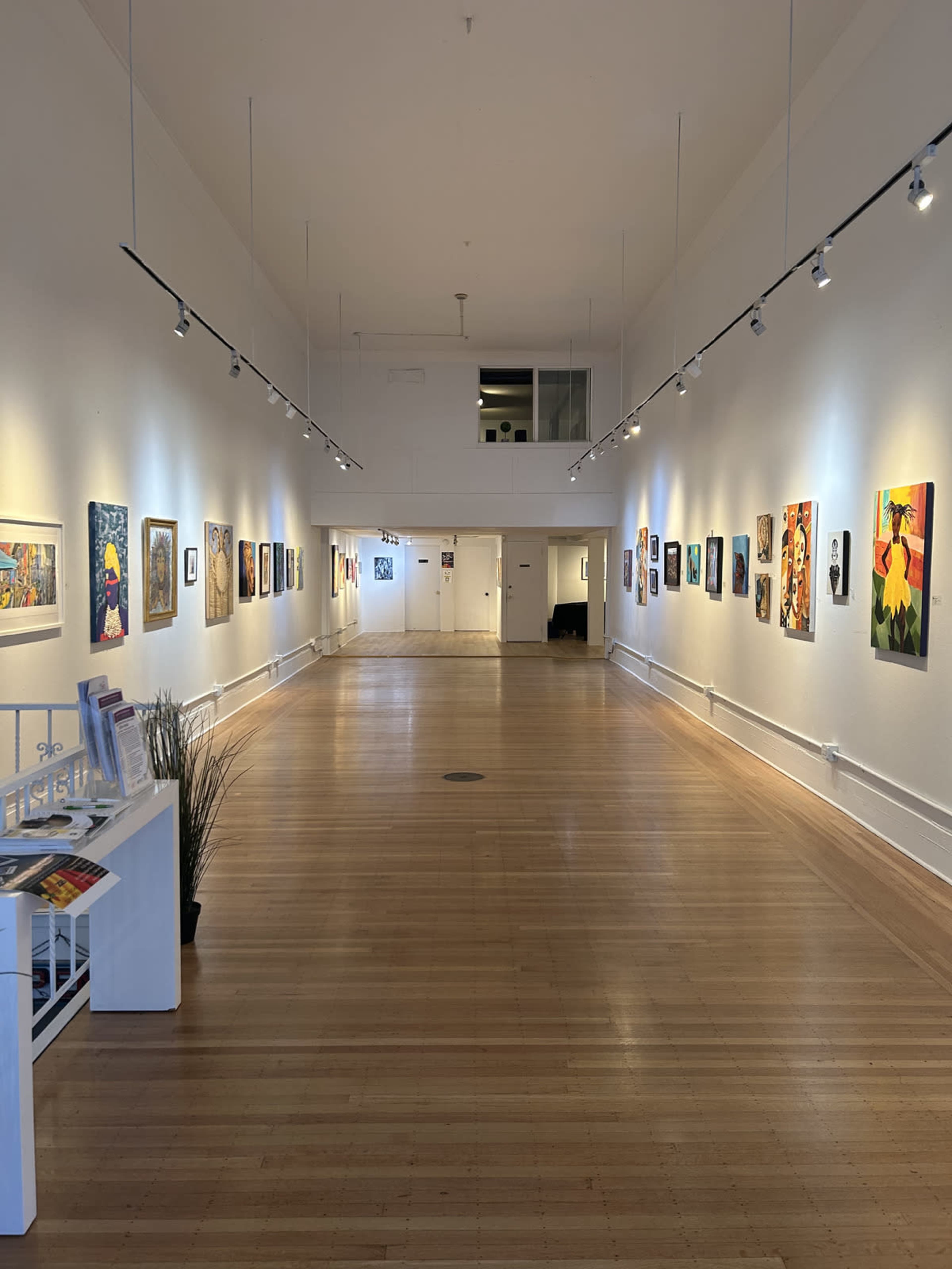The image shows a spacious art gallery with white walls, wooden floors, and a series of paintings hanging along the walls, illuminated by track lighting.
