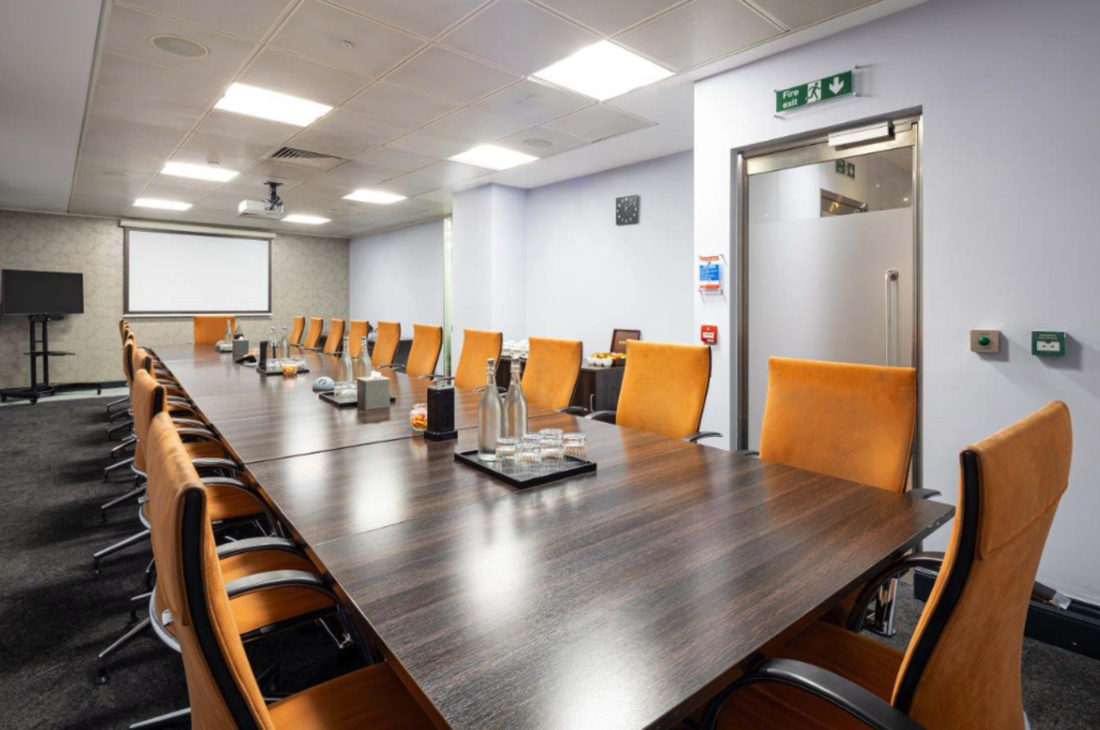 A large conference room features a long wooden table surrounded by orange chairs, with a projector, screen, and equipment for presentations.