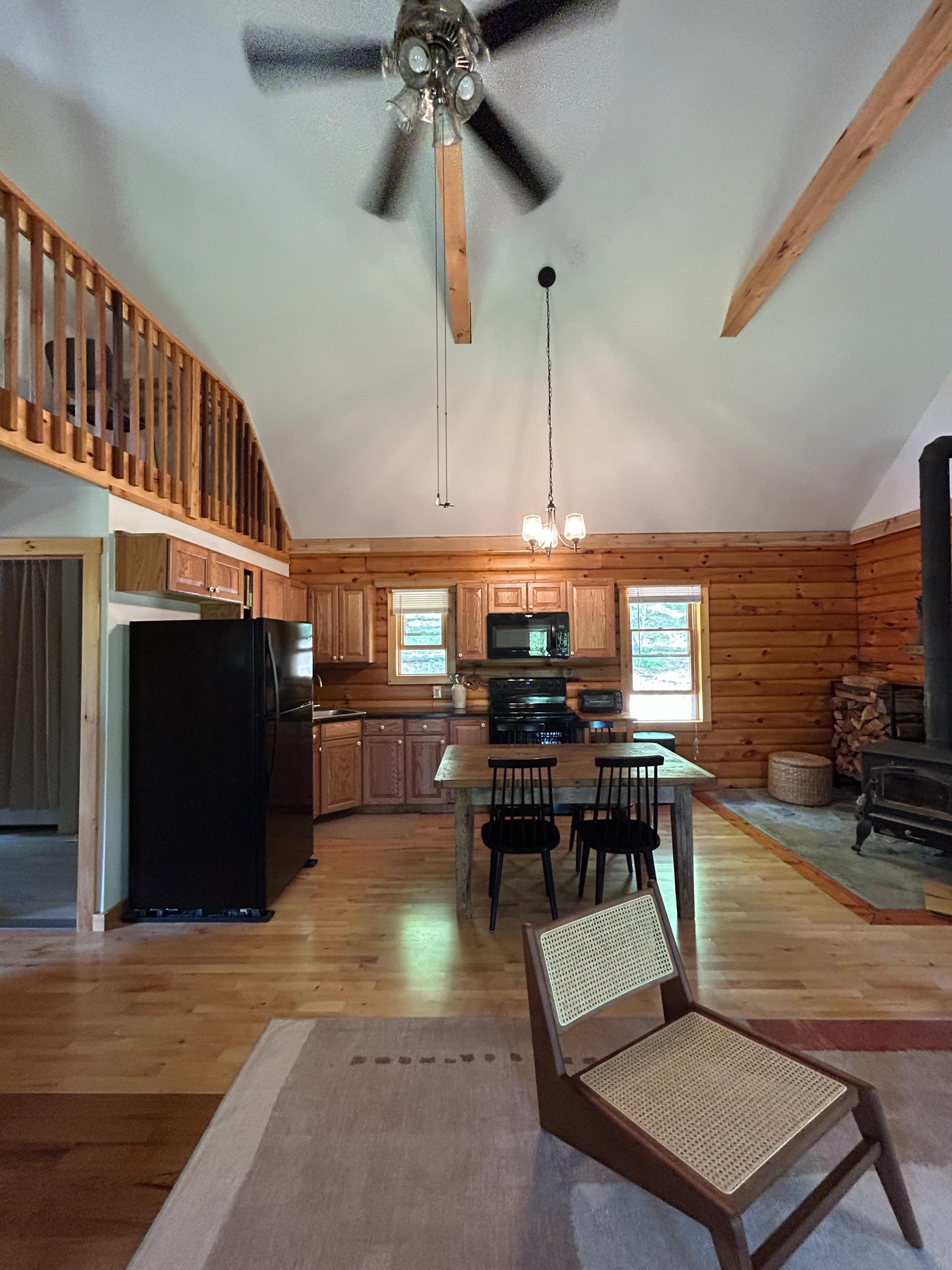 The image shows a cozy cabin interior featuring wooden walls, a kitchen area with dark cabinets and appliances, a dining table with two chairs, and a living space with a woven lounge chair and a wood stove.