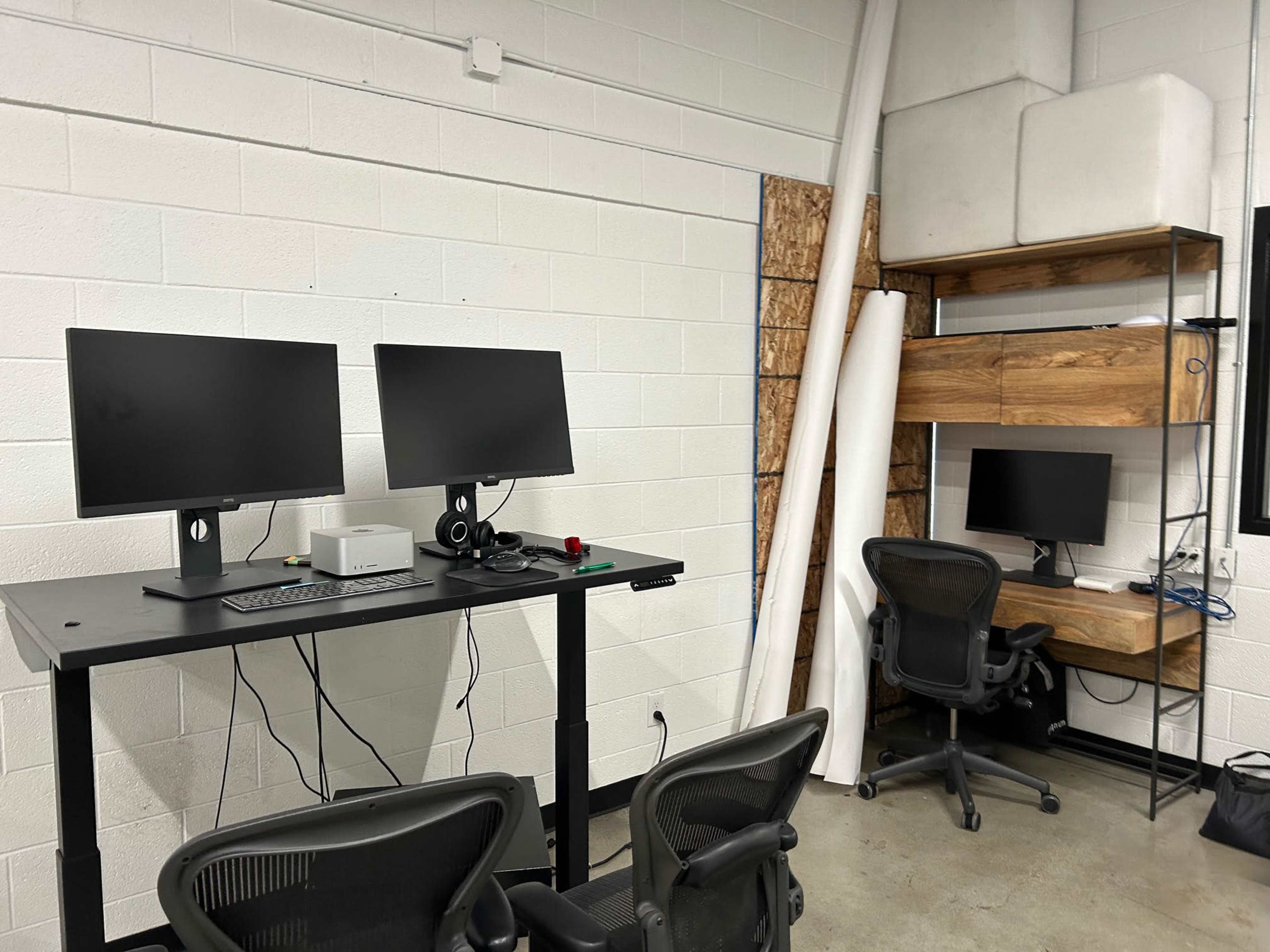 A workspace with two monitors on a black standing desk, a desktop computer, and a smaller monitor on a wooden shelf, with two office chairs.