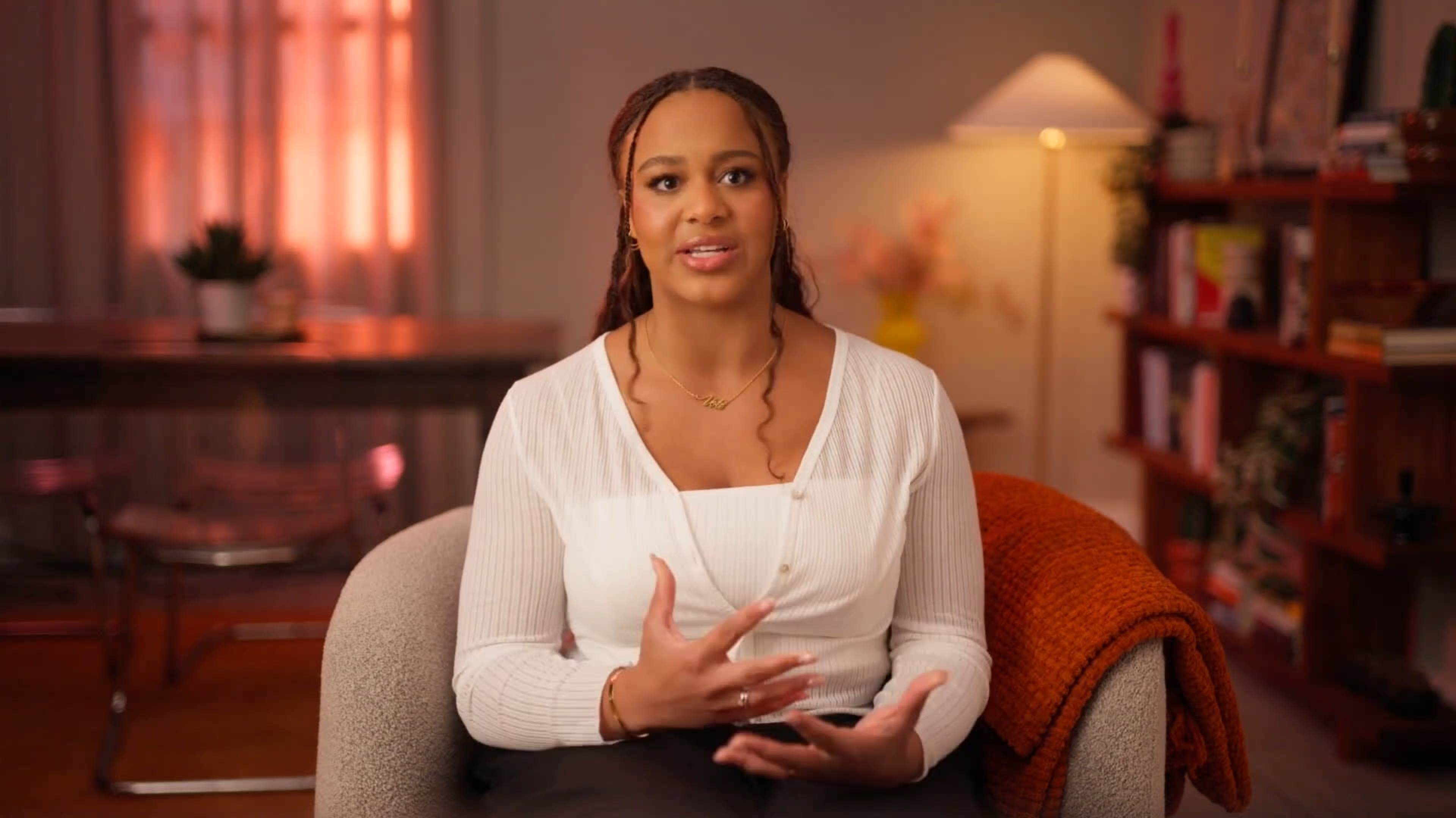 A woman with long braided hair is sitting in a cozy chair, speaking and gesturing while surrounded by warm lighting and decorative elements.