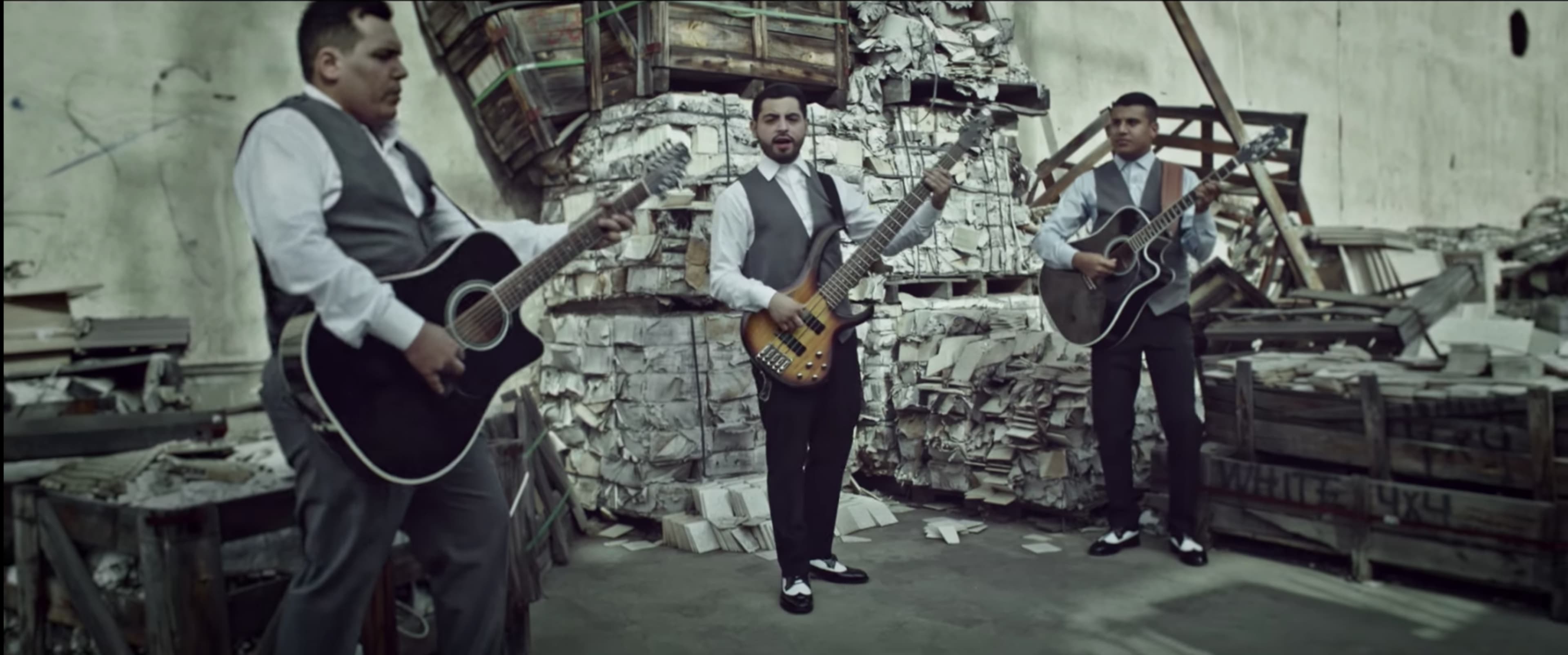 Three musicians in formal attire play guitars in a cluttered, industrial space filled with stacks of papers and wooden pallets.