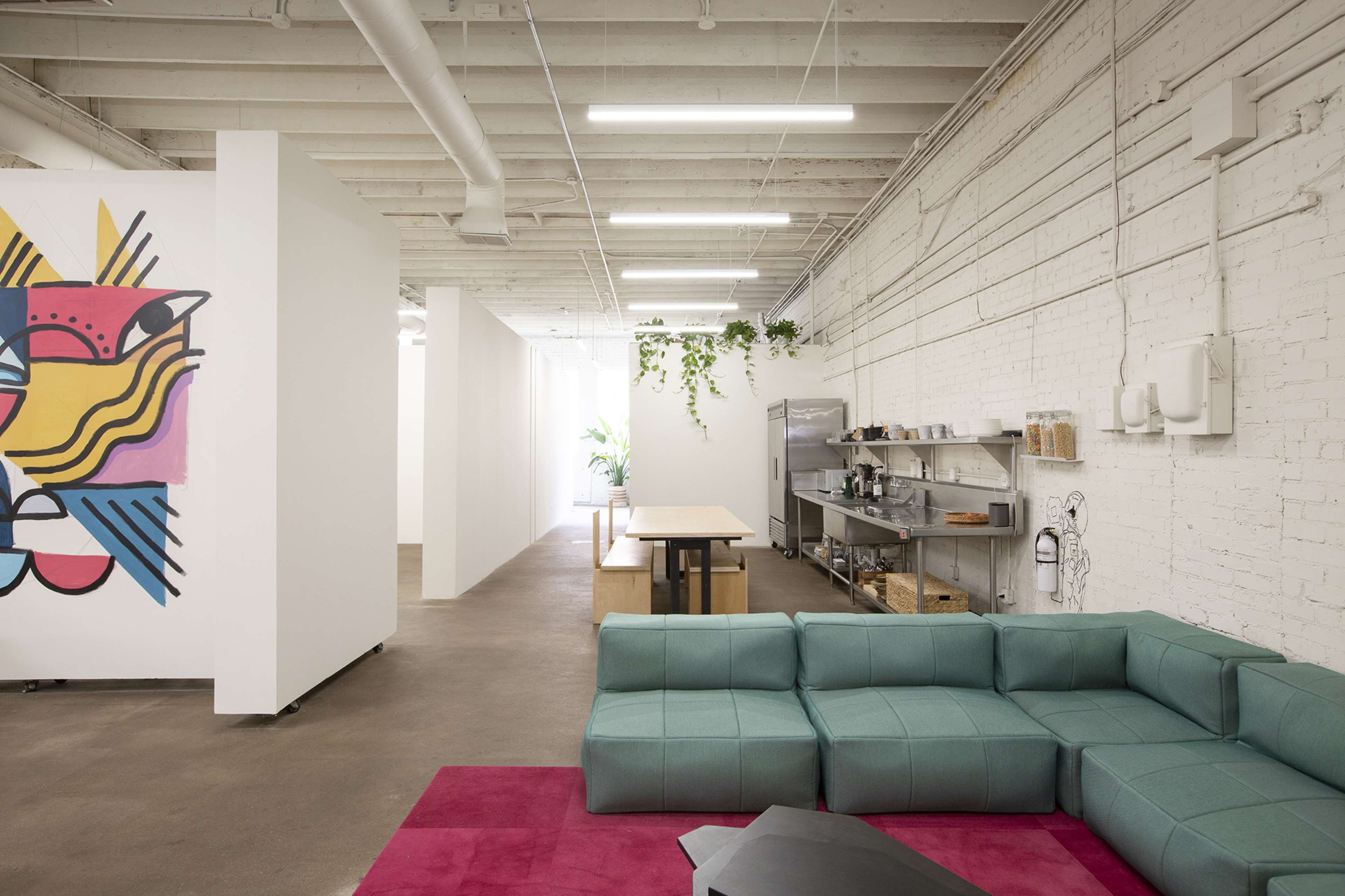 A modern office space featuring a turquoise sectional couch, a colorful mural on the wall, and an open kitchen area with light-colored furnishings.