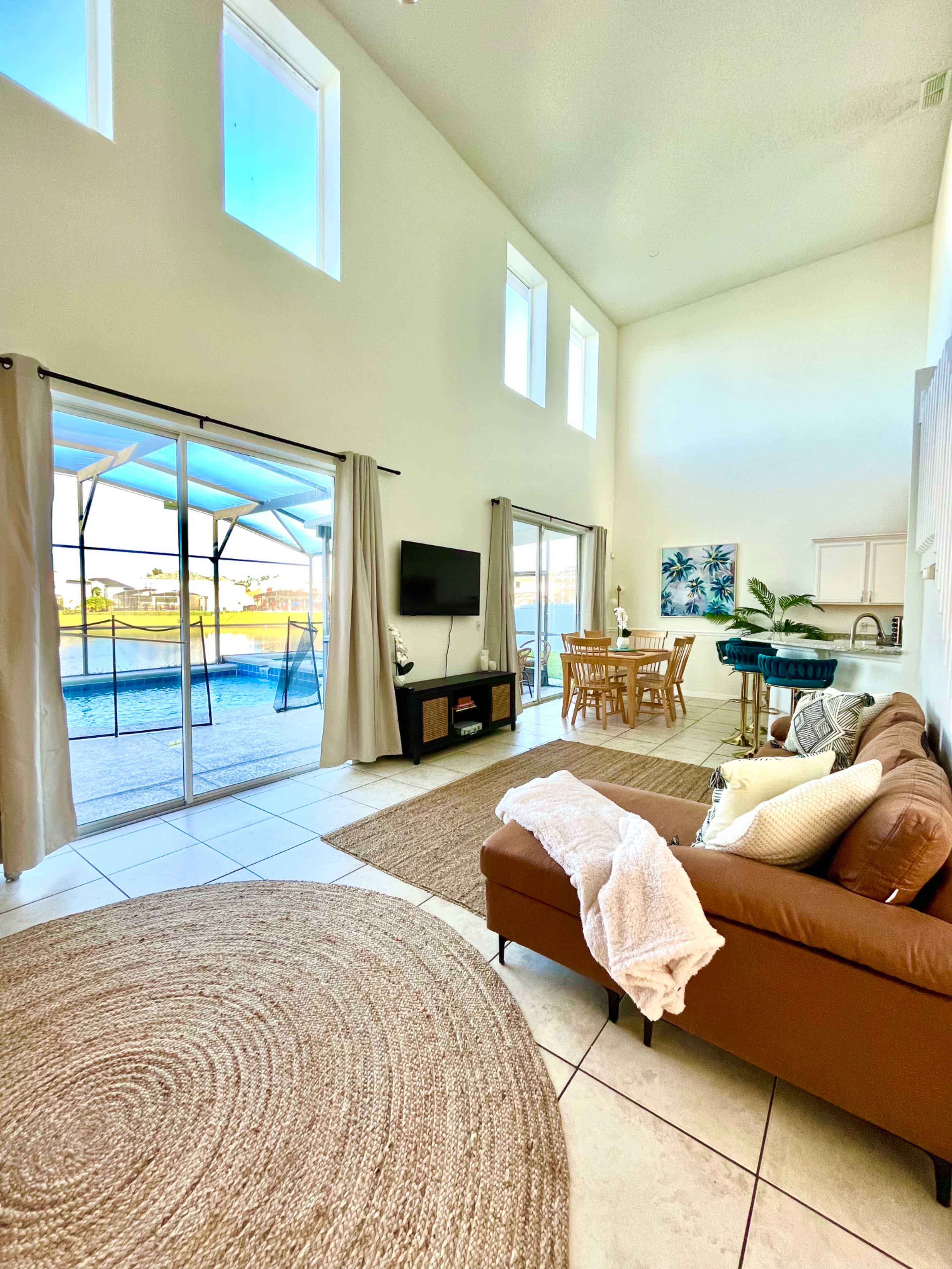 A spacious living room with a brown sofa, a round rug, a dining area, and large windows offering a view of an outdoor pool.