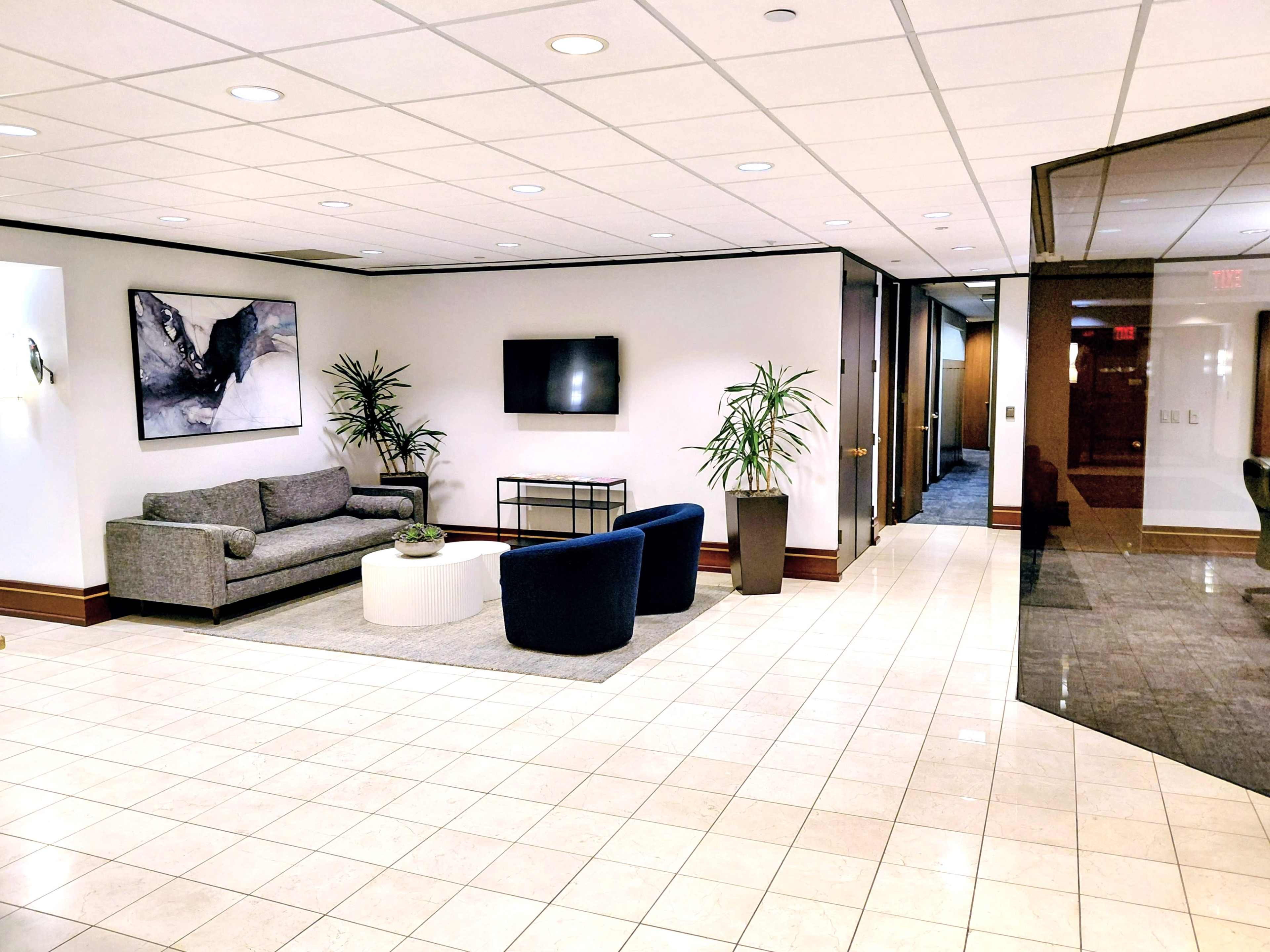 The image shows a modern lobby area featuring a gray sofa, two blue chairs, a white circular coffee table, and potted plants, along with a wall-mounted television and framed artwork in a well-lit space with tile flooring.