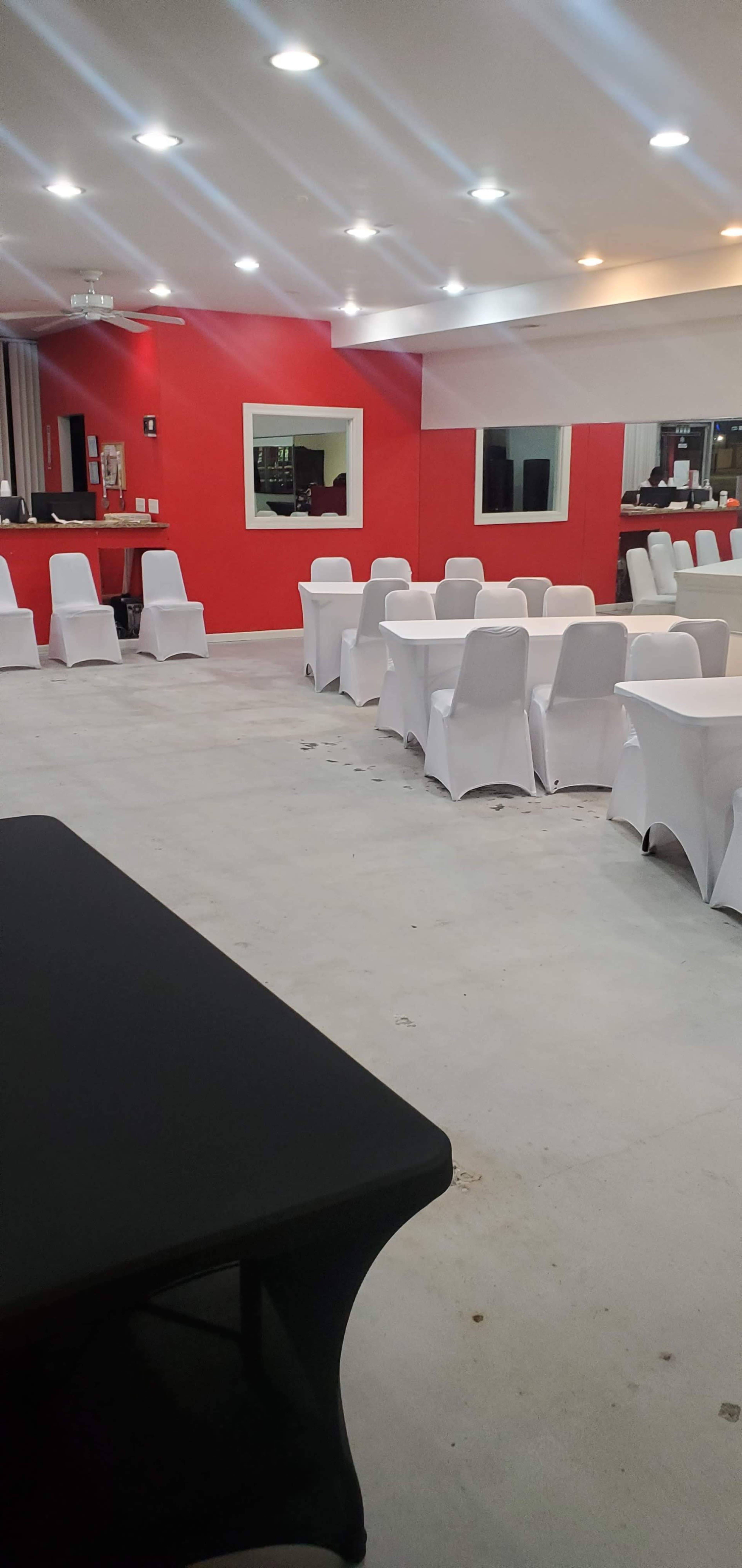The image shows a spacious event venue with white tables covered by tablecloths and a red-painted wall.