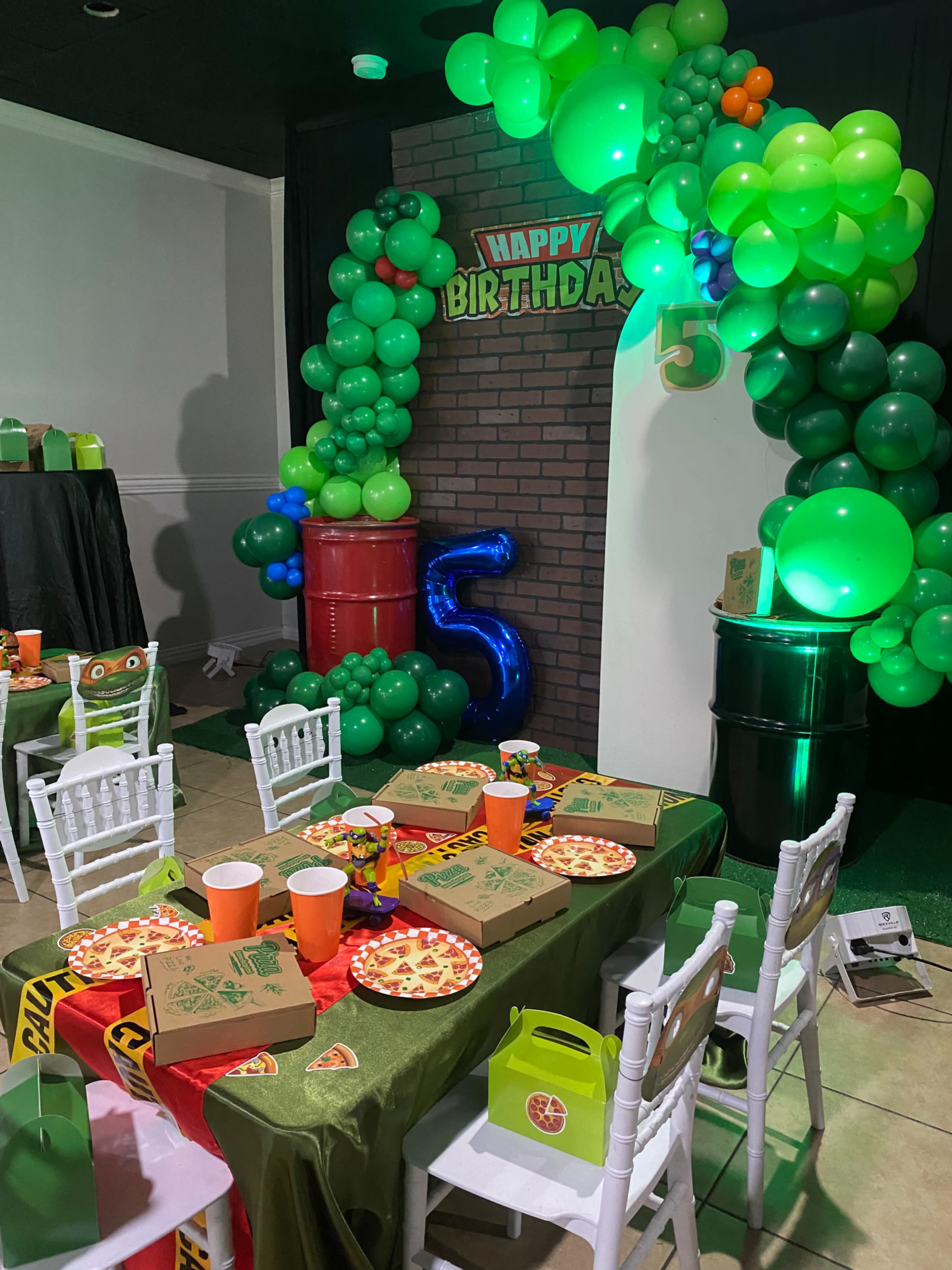 The image shows a colorful birthday party setup featuring a decorated table with pizza boxes, snacks, vibrant balloons, and a backdrop that includes a "Happy Birthday" sign and the number 5.