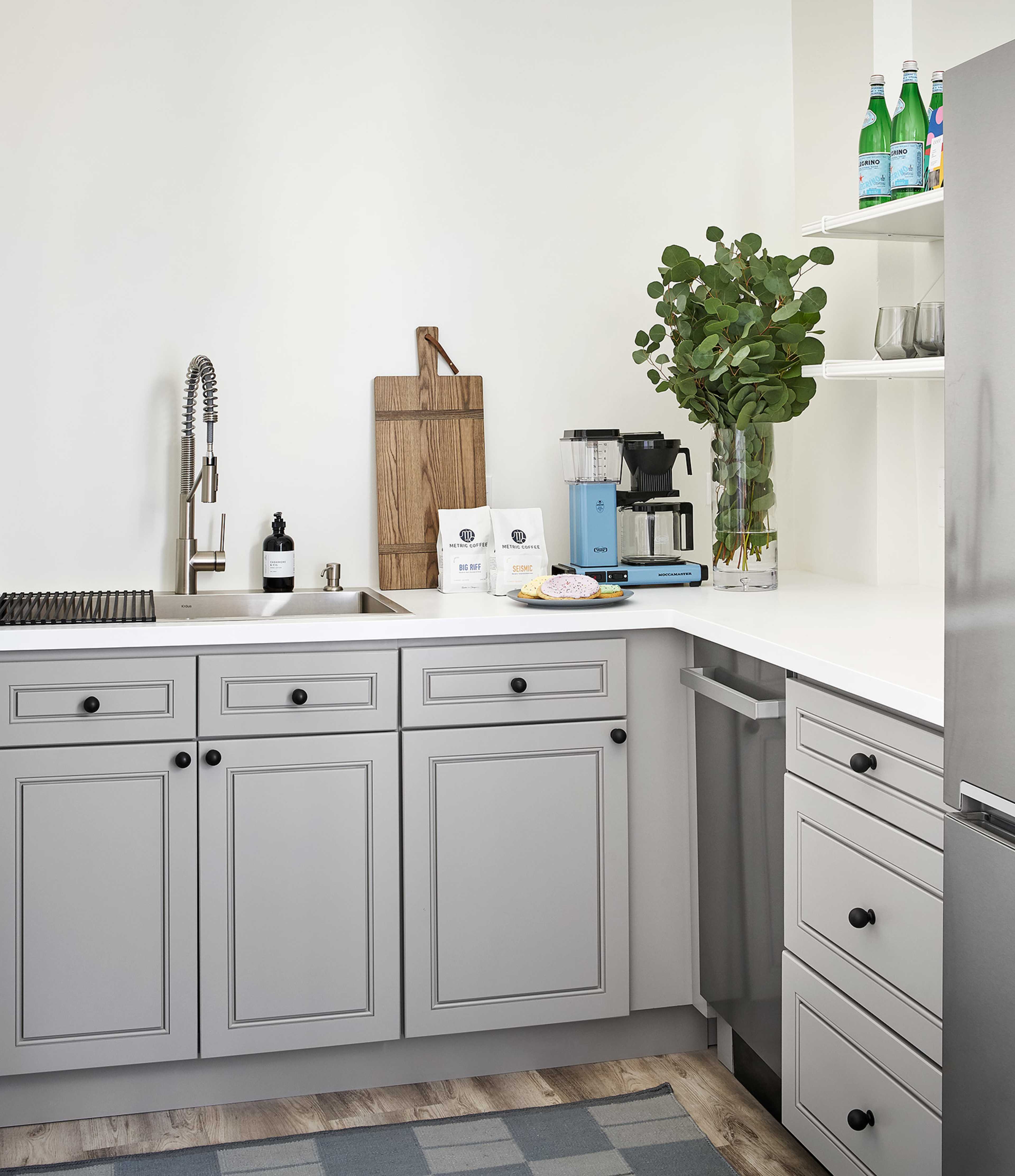 A modern kitchen features gray cabinetry, a stainless steel fridge, a sink with a sleek faucet, and a countertop displaying kitchen appliances and a cutting board.