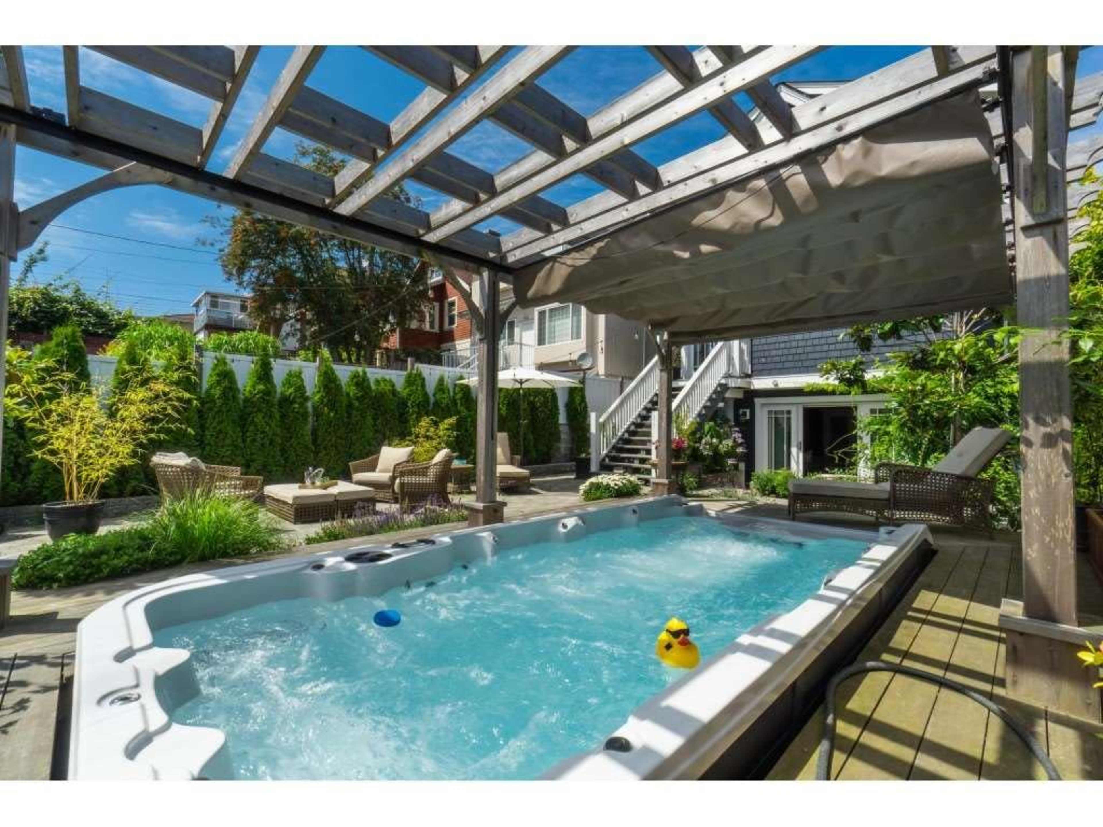 The image shows a spacious outdoor area featuring a hot tub surrounded by greenery, seating areas, and a wooden pergola.