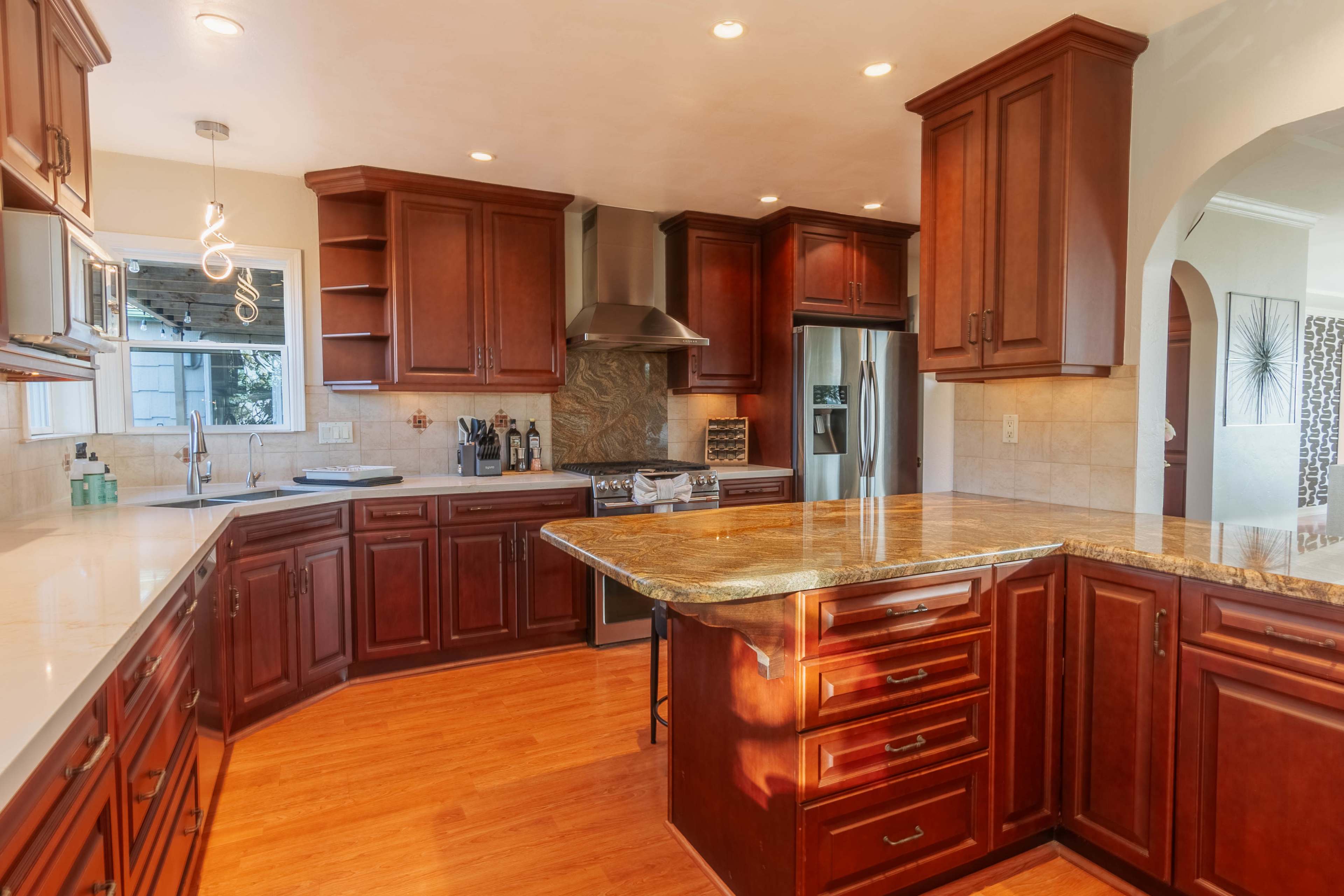 The image shows a spacious kitchen with dark wooden cabinets, a large island countertop, and modern appliances.