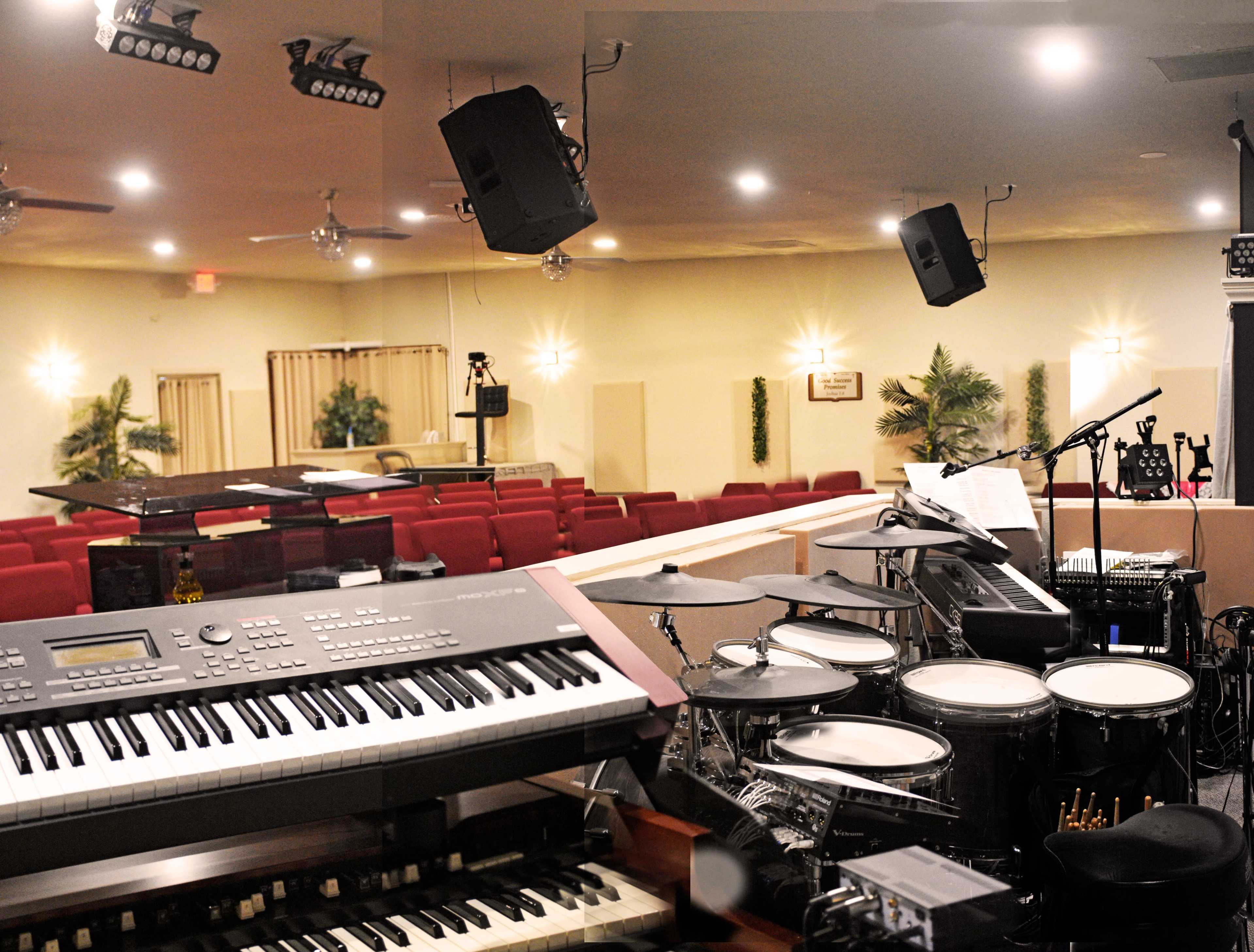 A music setup with electronic keyboards and a drum set positioned in front of an empty auditorium with red chairs.