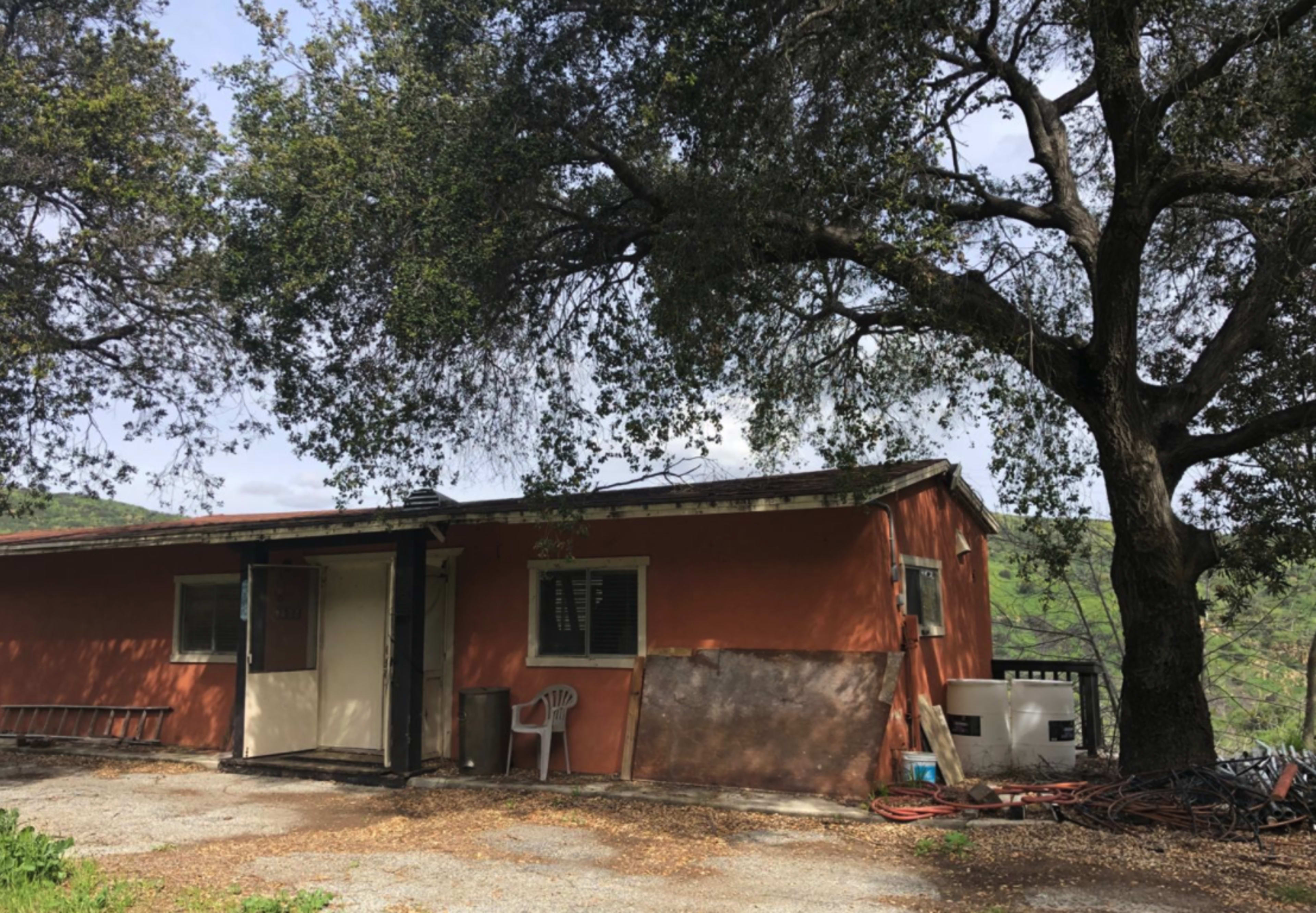 A single-story orange house with a porch is situated beneath a large tree, surrounded by gravel and greenery.