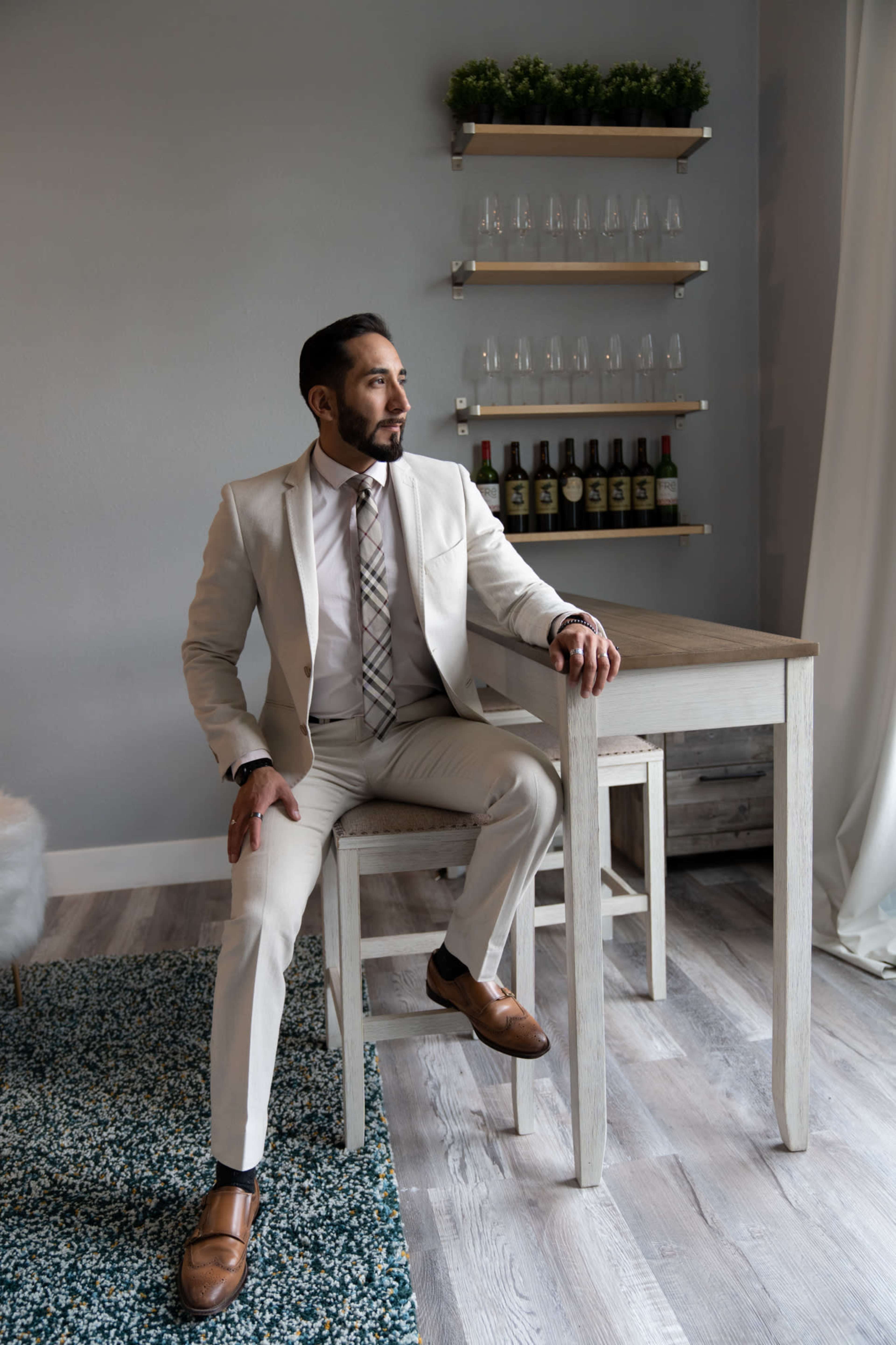 A man in a light-colored suit sits on a bar stool beside a table against a gray wall with shelves holding bottles and glasses.