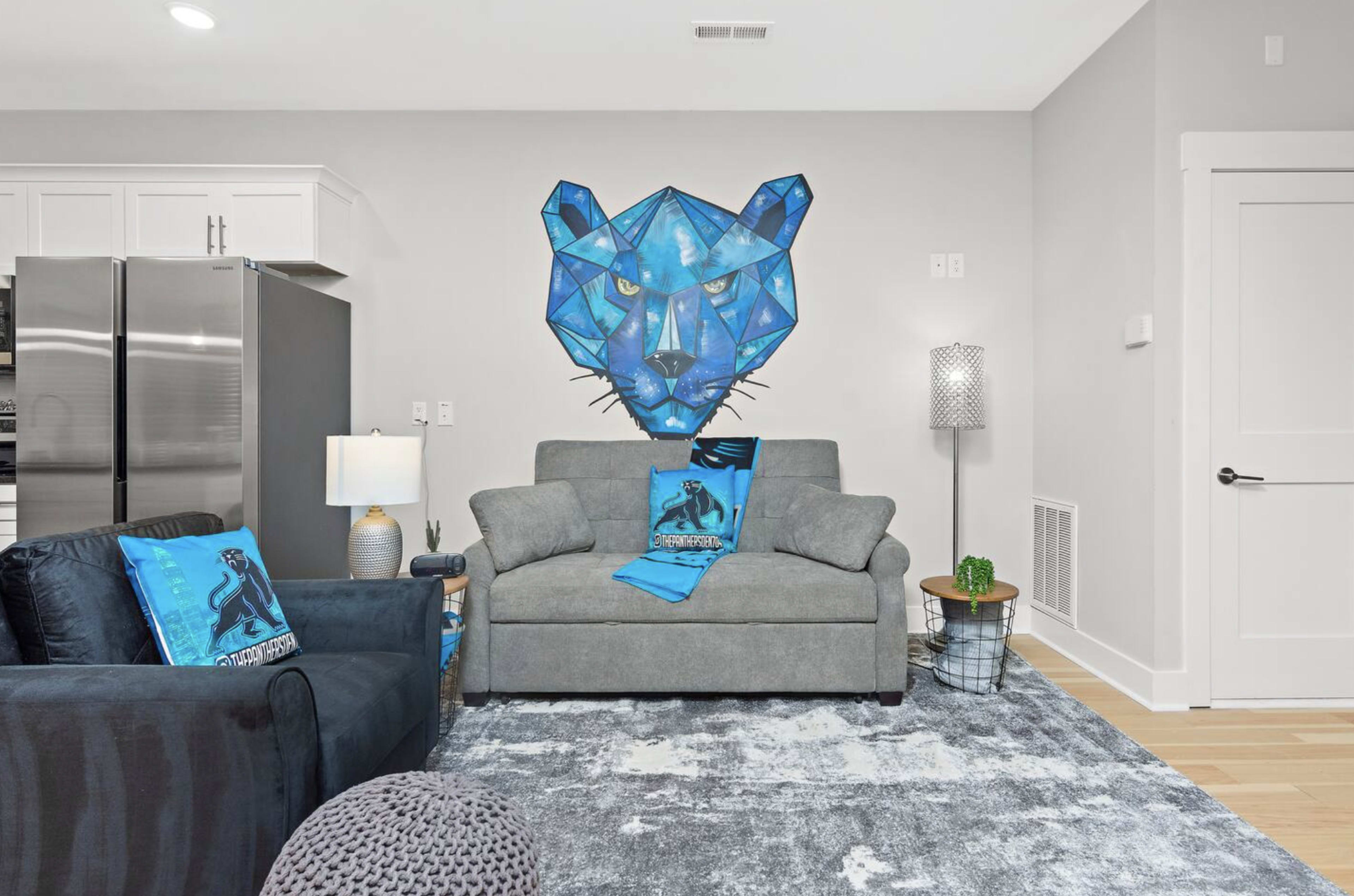A modern living room with a gray sofa, an accent chair, and a geometric blue cat mural on the wall behind them.