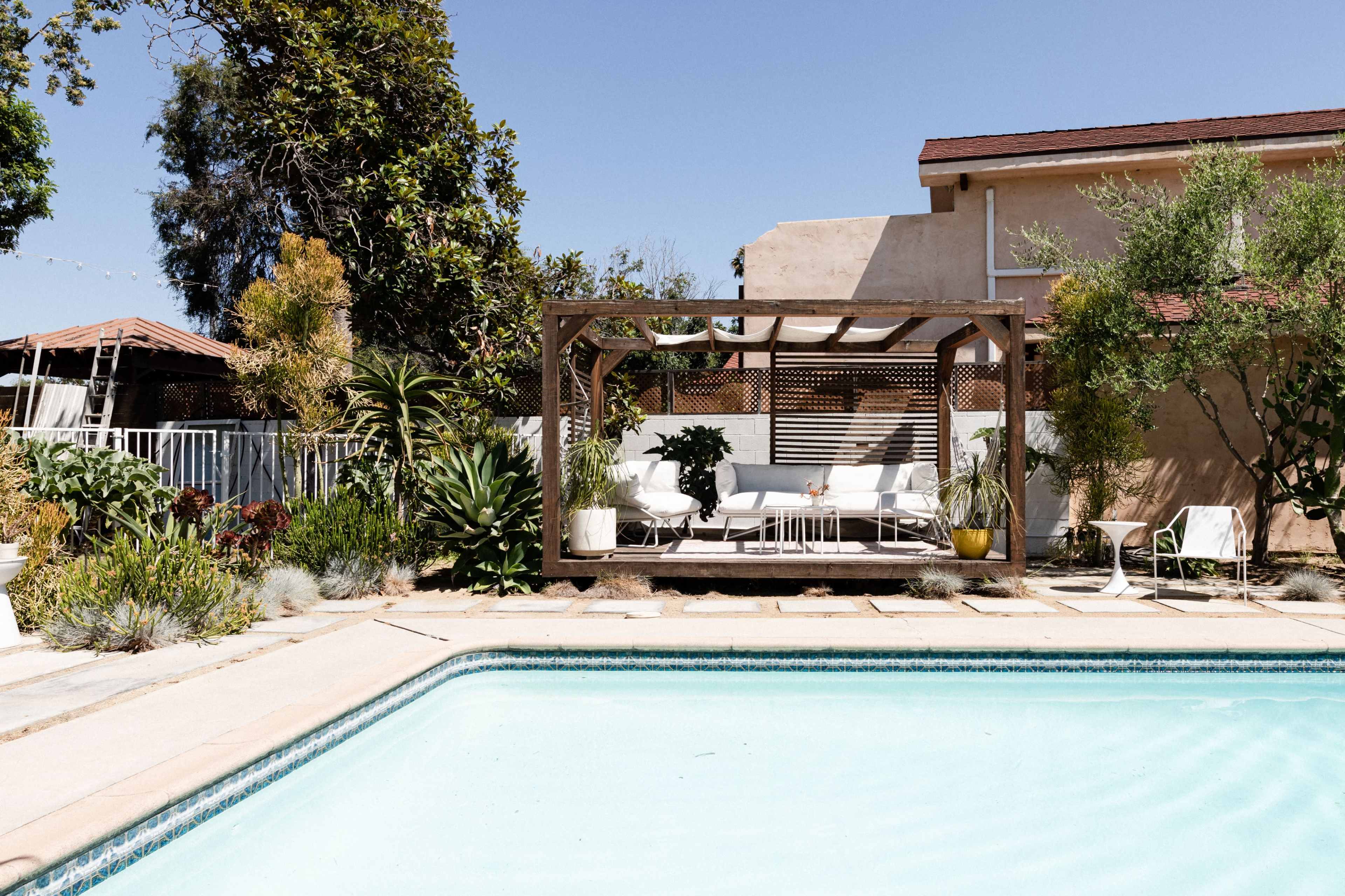 A swimming pool with a wooden pergola and seating is surrounded by plants and a residential building in a sunny outdoor setting.