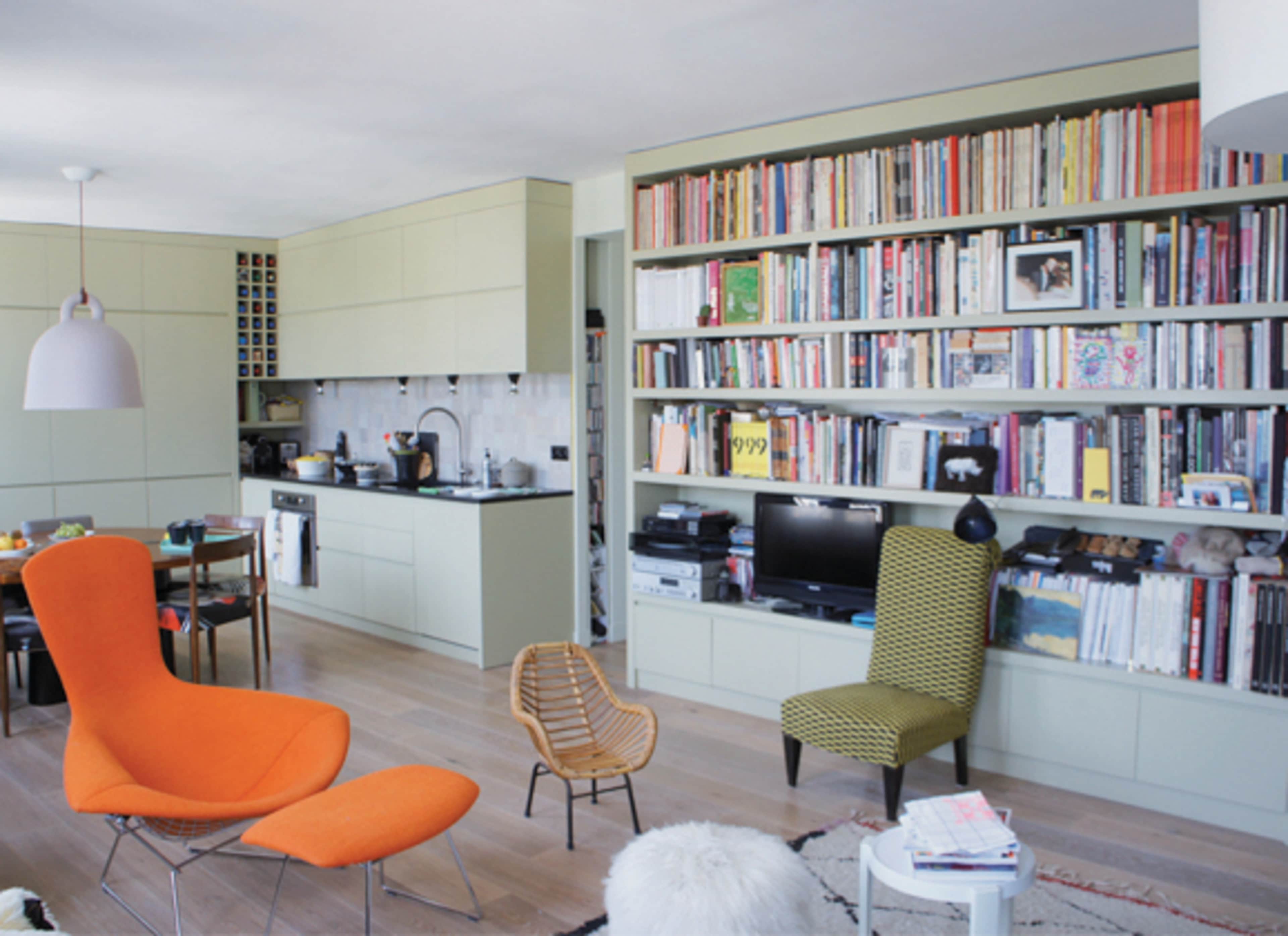 The image shows a modern living space featuring a kitchen area with green cabinetry, a large bookshelf filled with books, and colorful furniture, including an orange chair and a green patterned chair.
