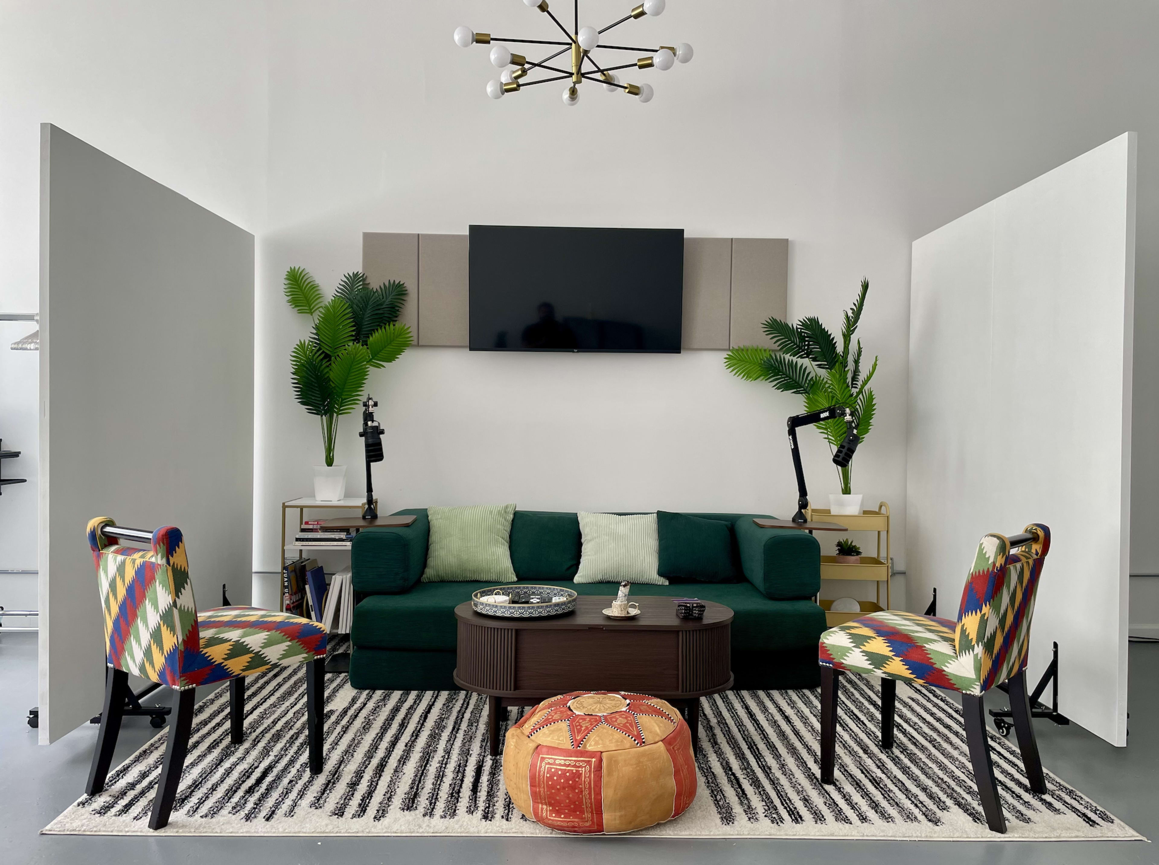 The image shows a modern living space featuring a green couch, a coffee table with decorative items, two patterned chairs, and a television mounted on a wall, framed by plants and partitions.