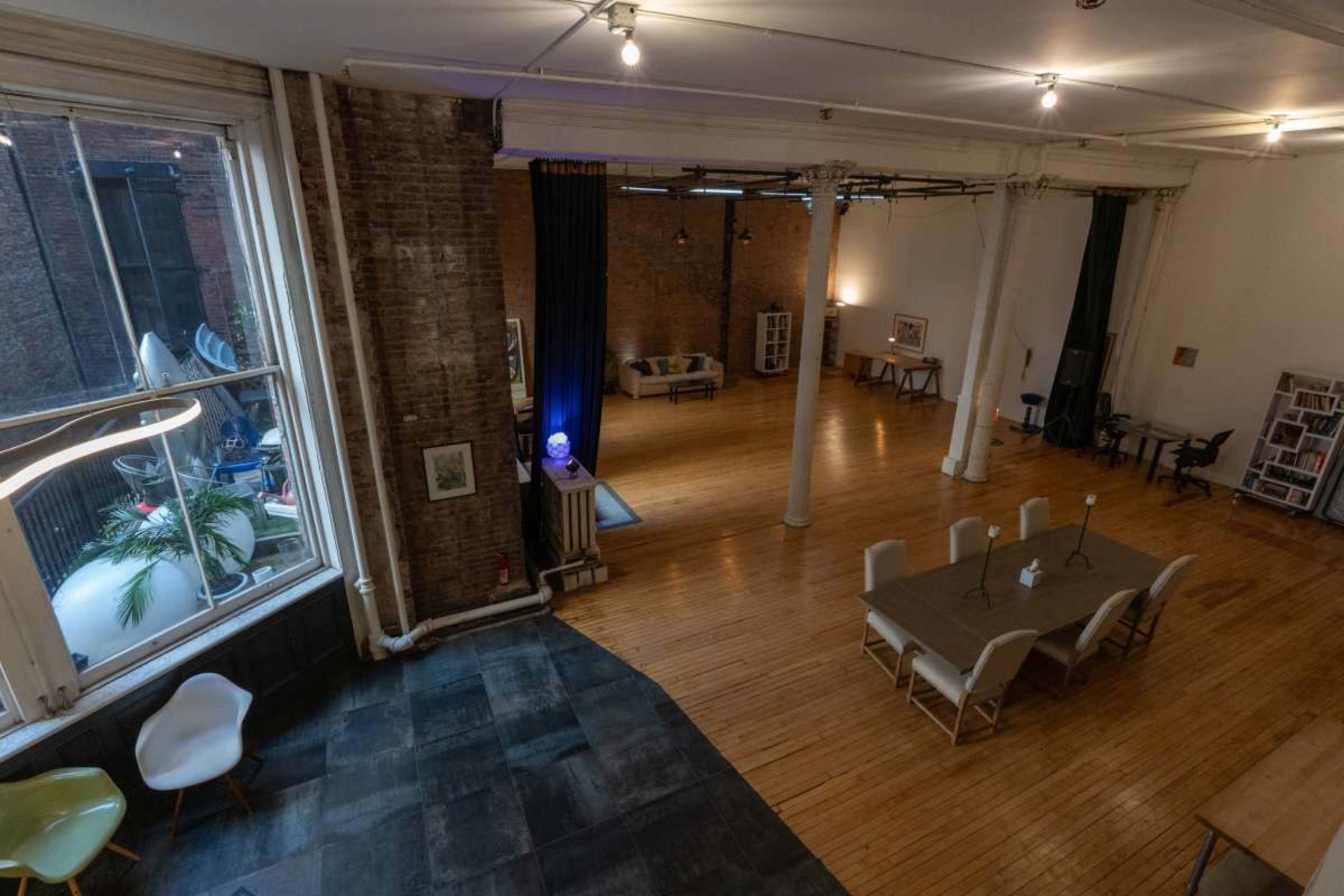 The image shows a spacious, well-lit open floor plan with wooden flooring, exposed brick walls, and distinct areas for seating and dining.